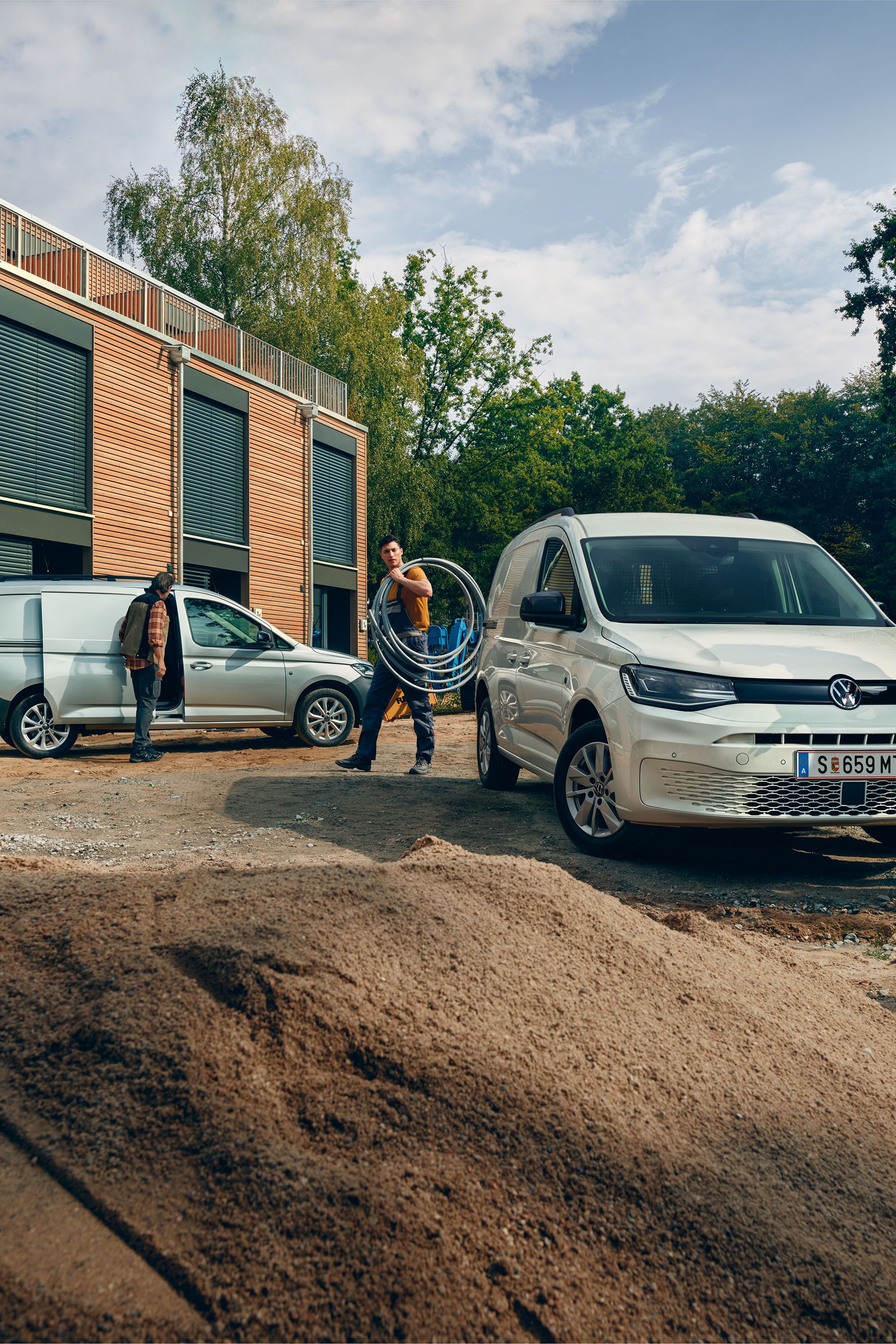 Два товарни автомобила Volkswagen Caddy на строителен обект, с трима професионалисти, носещи инструменти и оборудване.