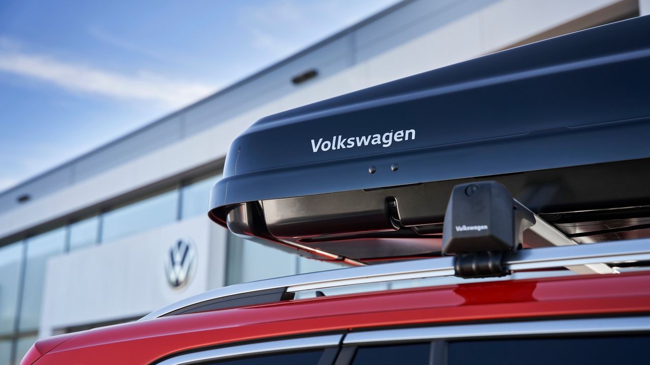 Червен автомобил Volkswagen е паркиран пред модерна сграда на фона на синьо небе. На покрива на автомобила е монтиран черен багажник с надпис "Volkswagen".