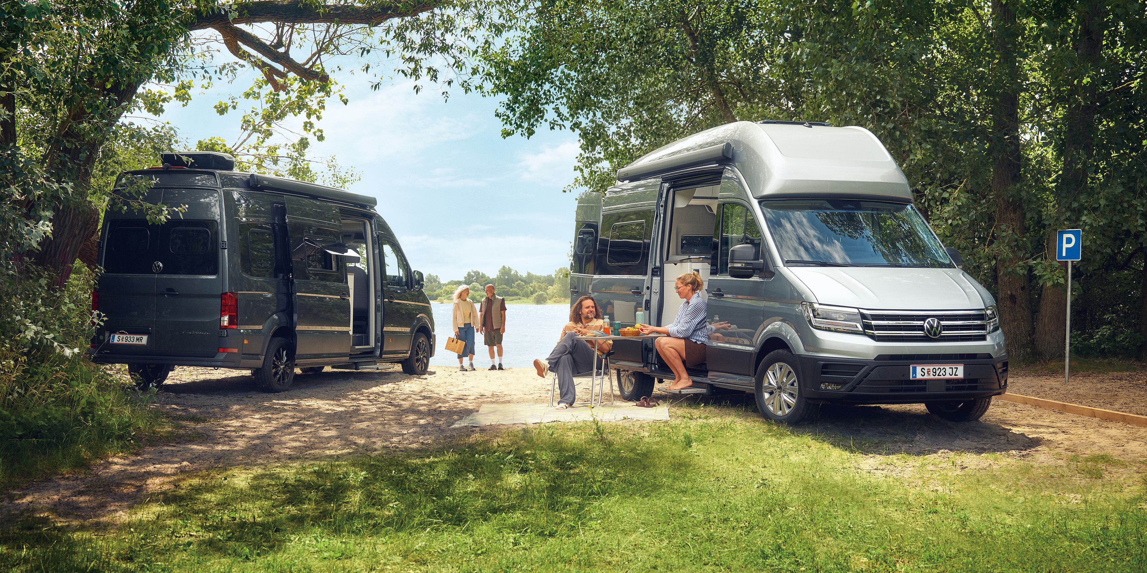 Zwei VW Grand California stehen auf einer grasbewachsenen Fläche in der Nähe eines Sees. Es gibt mehrere Personen; eine sitzt an einem Picknicktisch und zwei weitere gehen auf das Wasser zu. Die Szene ist von Bäumen und Grün umgeben.
