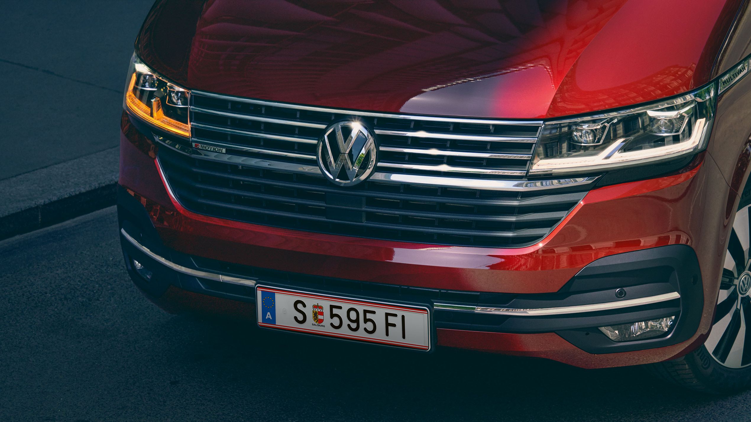 Червен автомобил с марка Volkswagen Multivan е паркиран на улица, като част от предната му част е осветена от слънчева светлина. Табелата му е със сериен номер SE595FI.