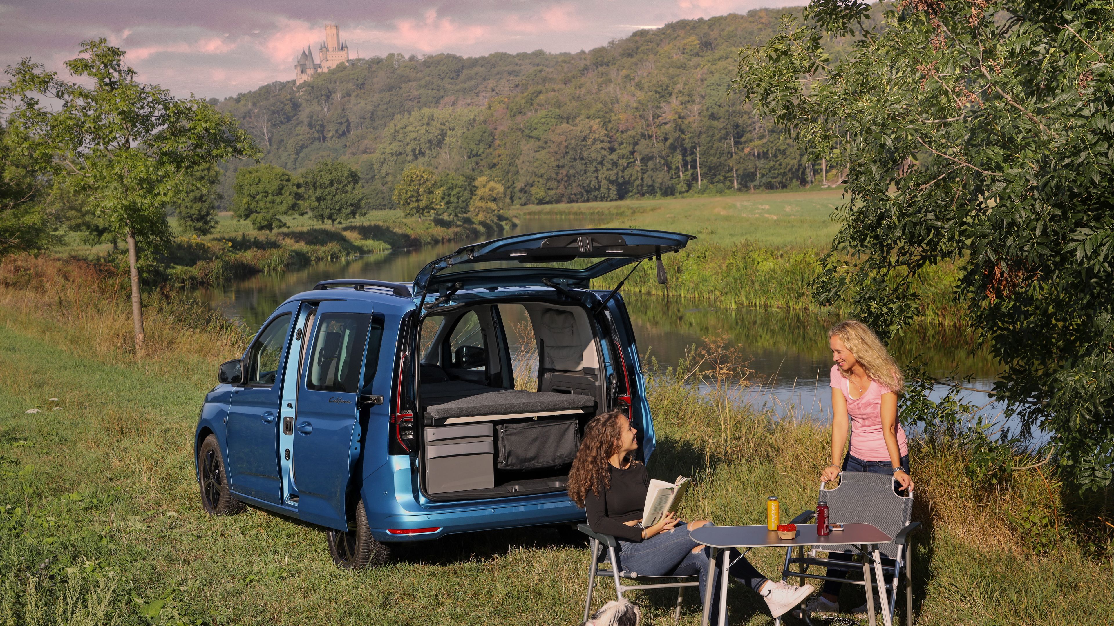 Blauer VW Caddy California mit Campingmöbel steht an See.