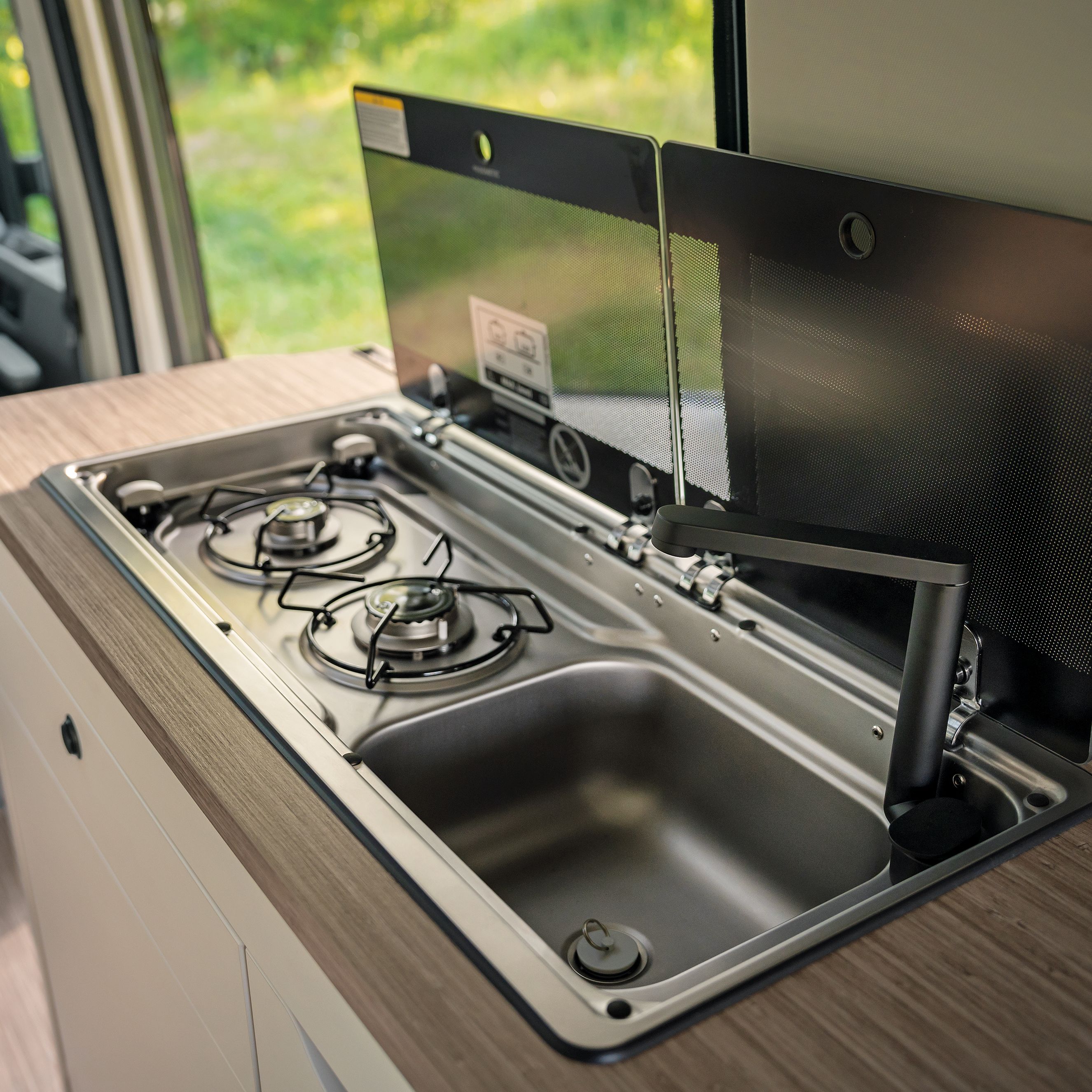 Das Bild zeigt eine kompakte Küchenzeile in einem Wohnmobil. Es sind zwei Gasherde und eine Spüle eingebaut. Die Rückwand ist hochgeklappt, wodurch die Kochflächen besser sichtbar sind.
