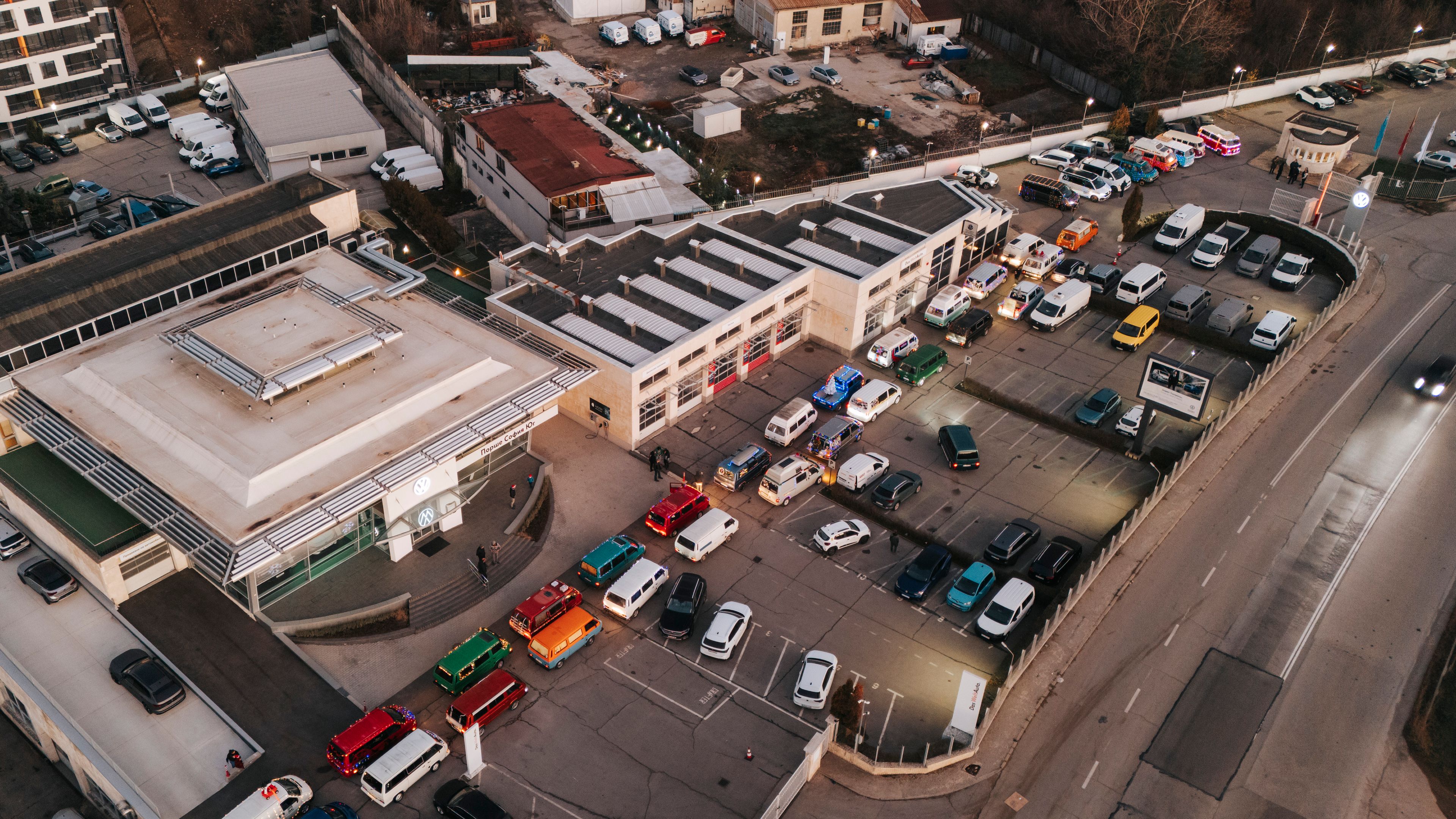 Въздушен изглед на Volkswagen  автомобилен паркинг, пълен с различни превозни средства, разположен до сградите на търговски обект. Път минава покрай паркинга, като няколко коли се движат по него. Виждат се офис сгради и паркинг с множество автомобили.