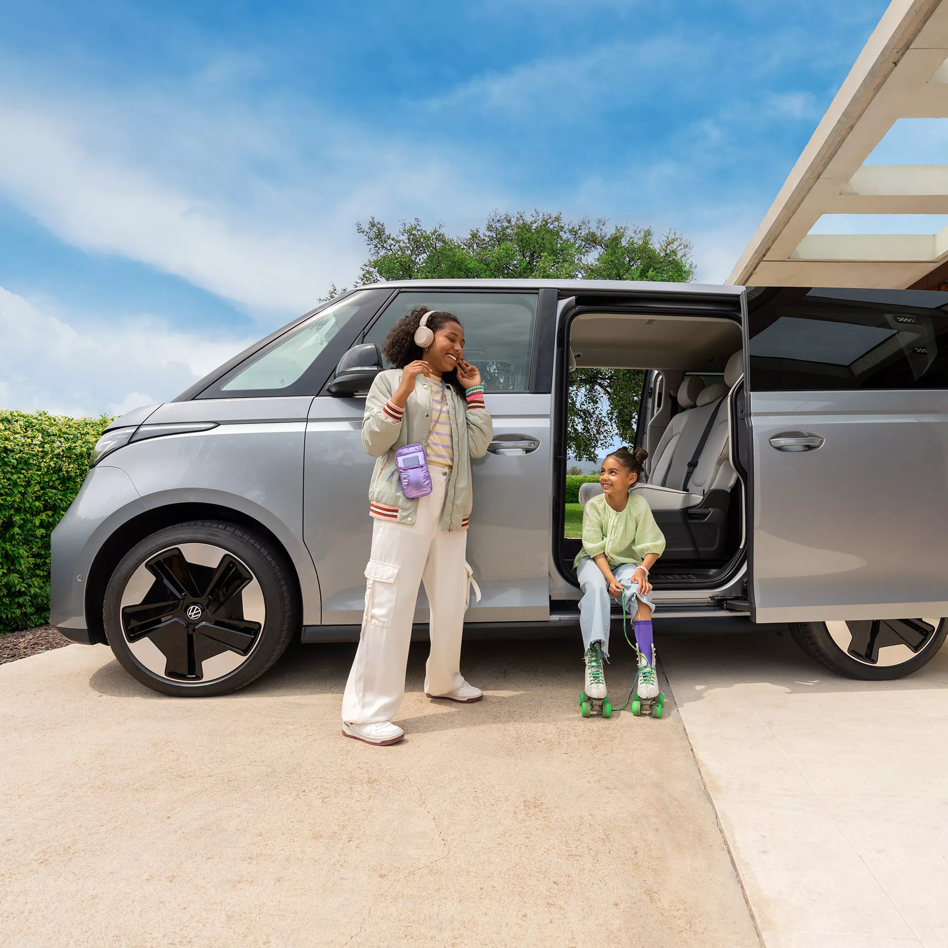 Две момичета се забавляват до сребрист Volkswagen ID. Buzz – едната със слушалки, другата с ролери, до отворената странична врата на автомобила.