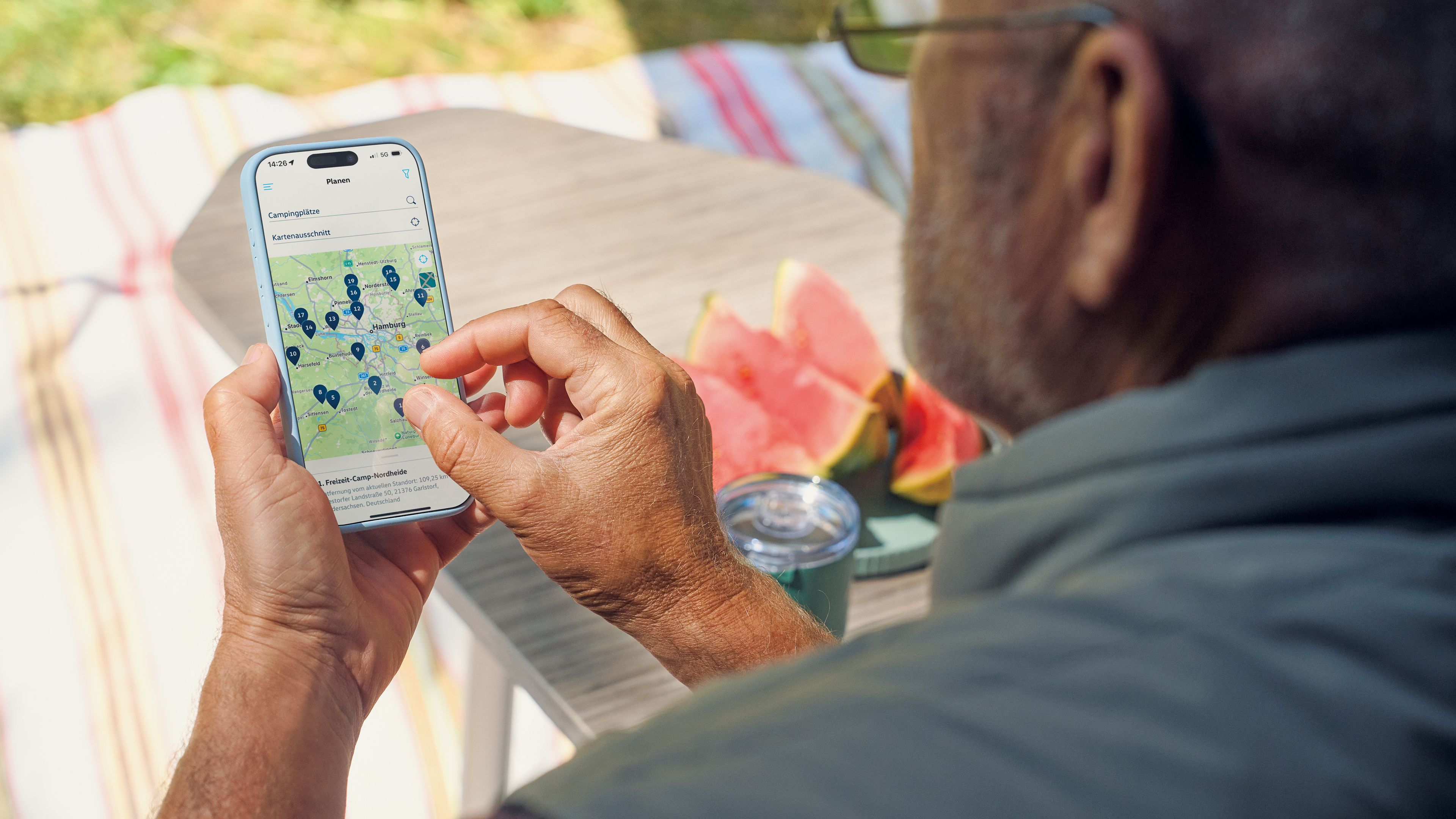 Ein älterer Mann nutzt ein Smartphone, um eine Karte mit mehreren Markierungen darauf anzusehen. Im Hintergrund sieht man einen Tisch mit Wassermelonenstücken und einen Streifenteppich.