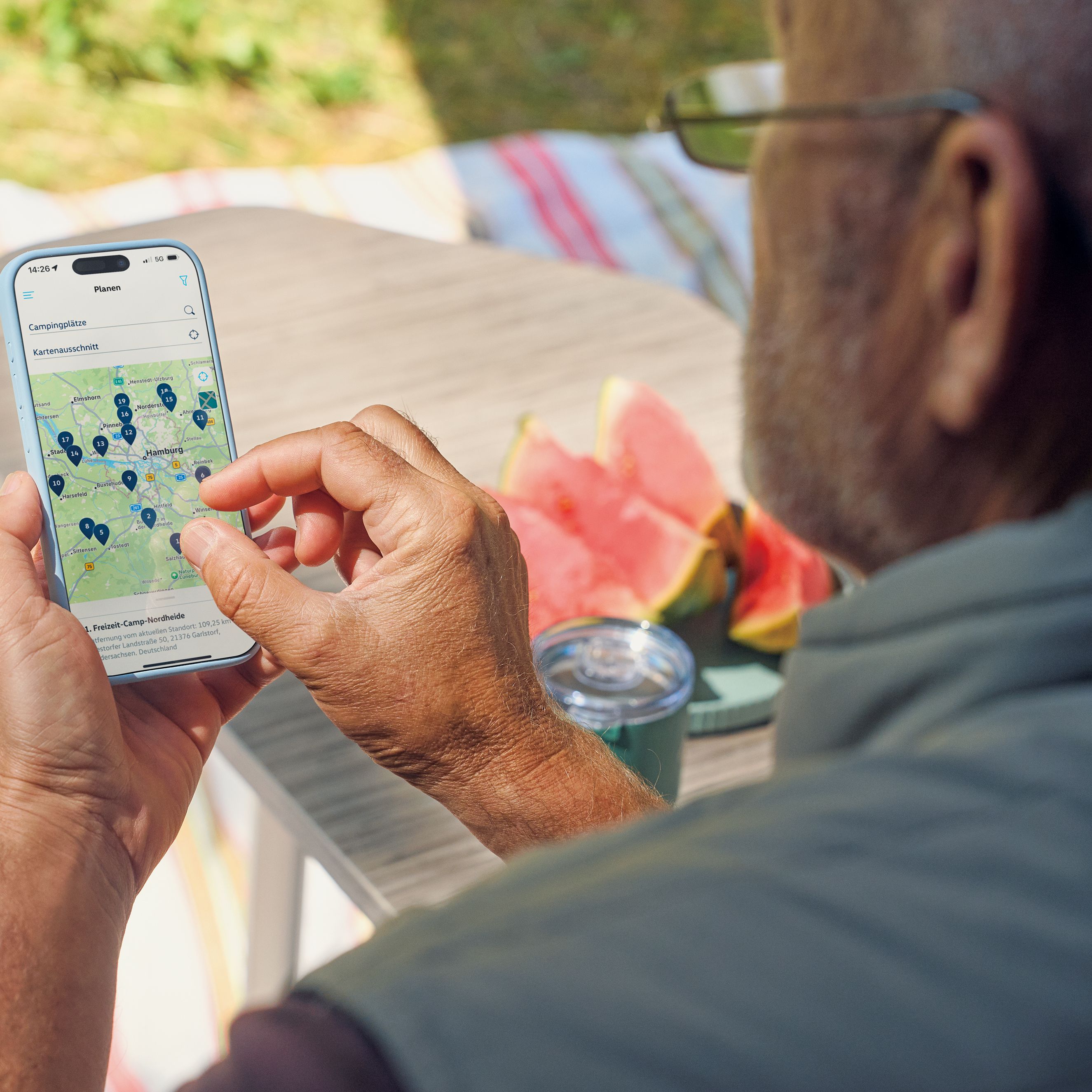 Ein älterer Mann nutzt ein Smartphone, um eine Karte mit mehreren Markierungen darauf anzusehen. Im Hintergrund sieht man einen Tisch mit Wassermelonenstücken und einen Streifenteppich.