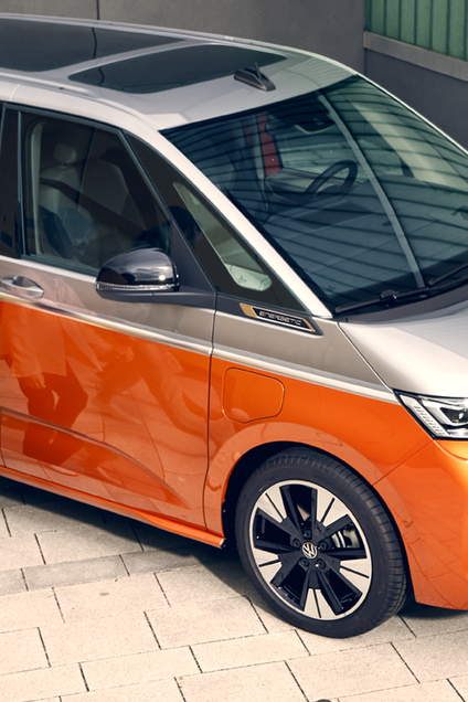 Щастливо семейство с деца пред оранжев Volkswagen Multivan, паркиран до сграда с електрическо зарядно.