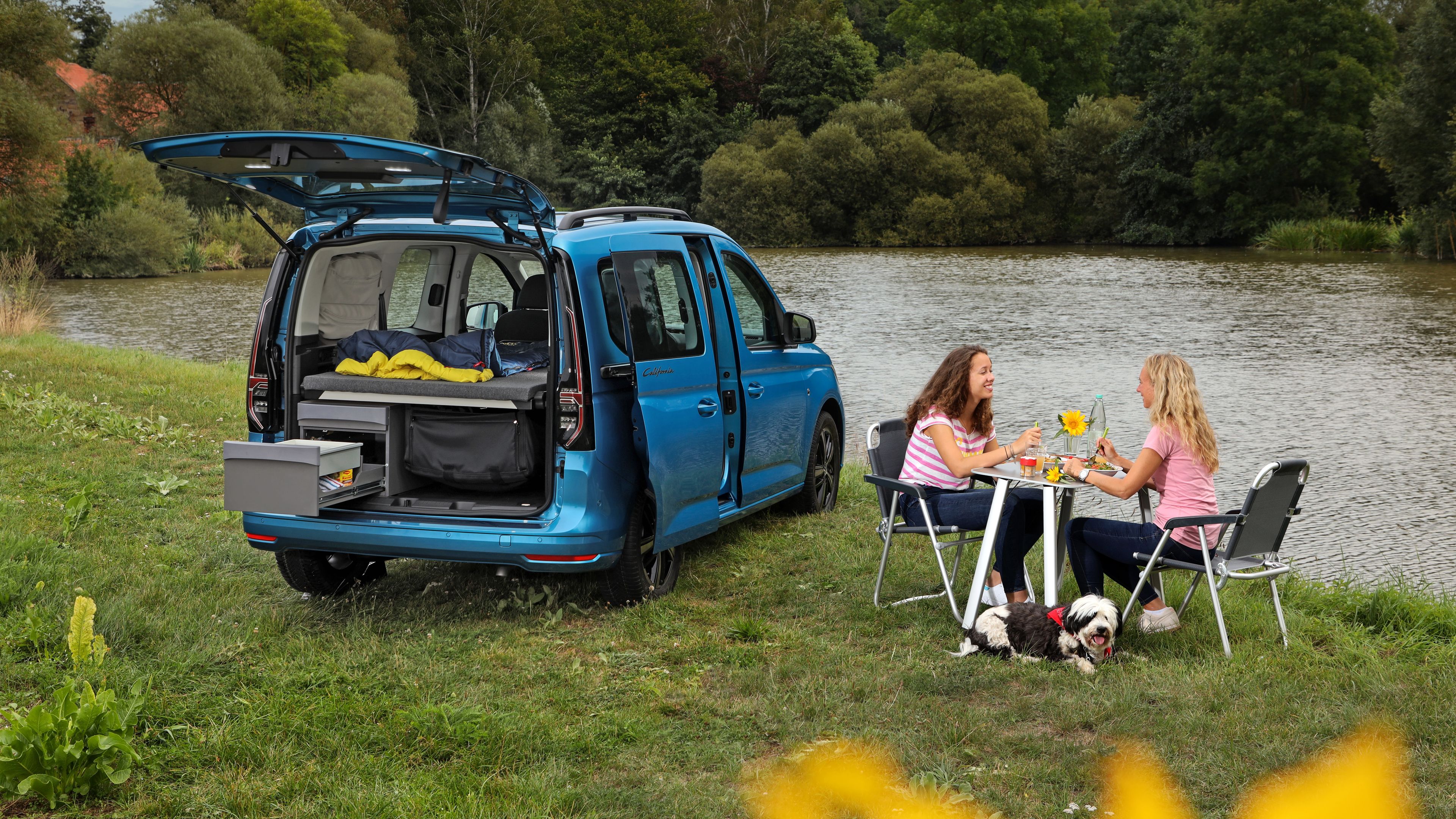 Blauer VW Caddy California steht an See und Frauen essen auf Campingmöbeln.