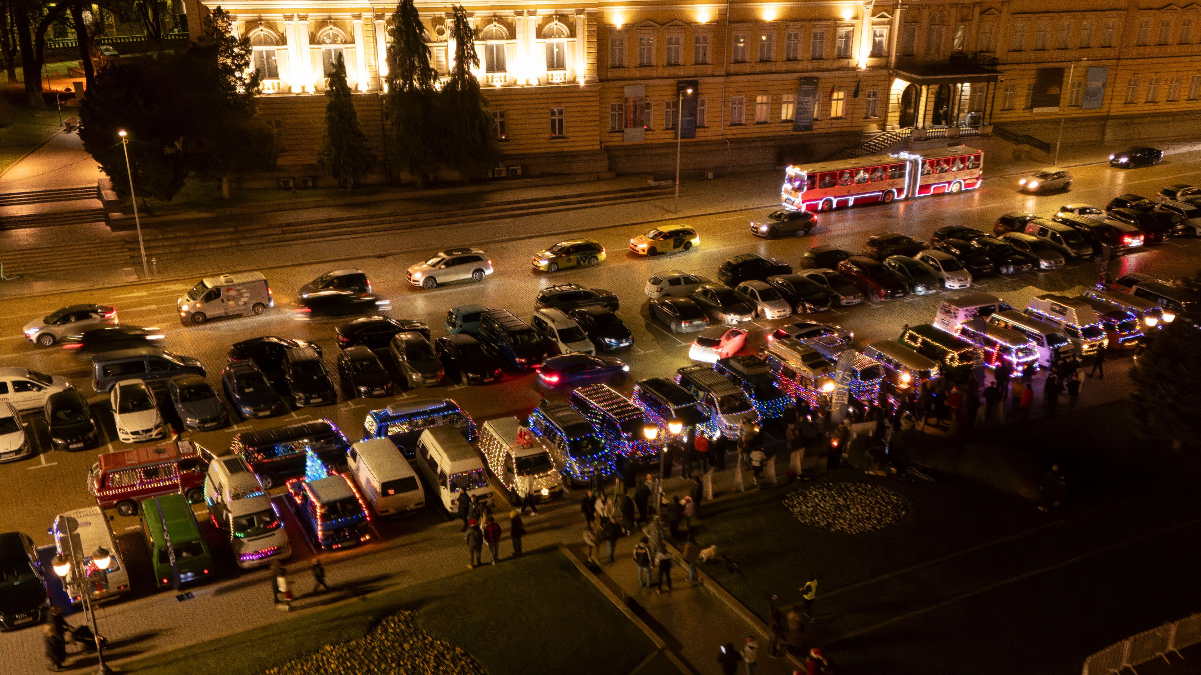 Вечерна сцена с паркирани Volkswagen автомобили, украсени с разноцветни светлини за празник. Отстрани има много хора, които се събират и разглеждат автомобила. В далечината се виждат автомобилите, движещи се по улицата, а в горната част на изображението - осветена сграда.