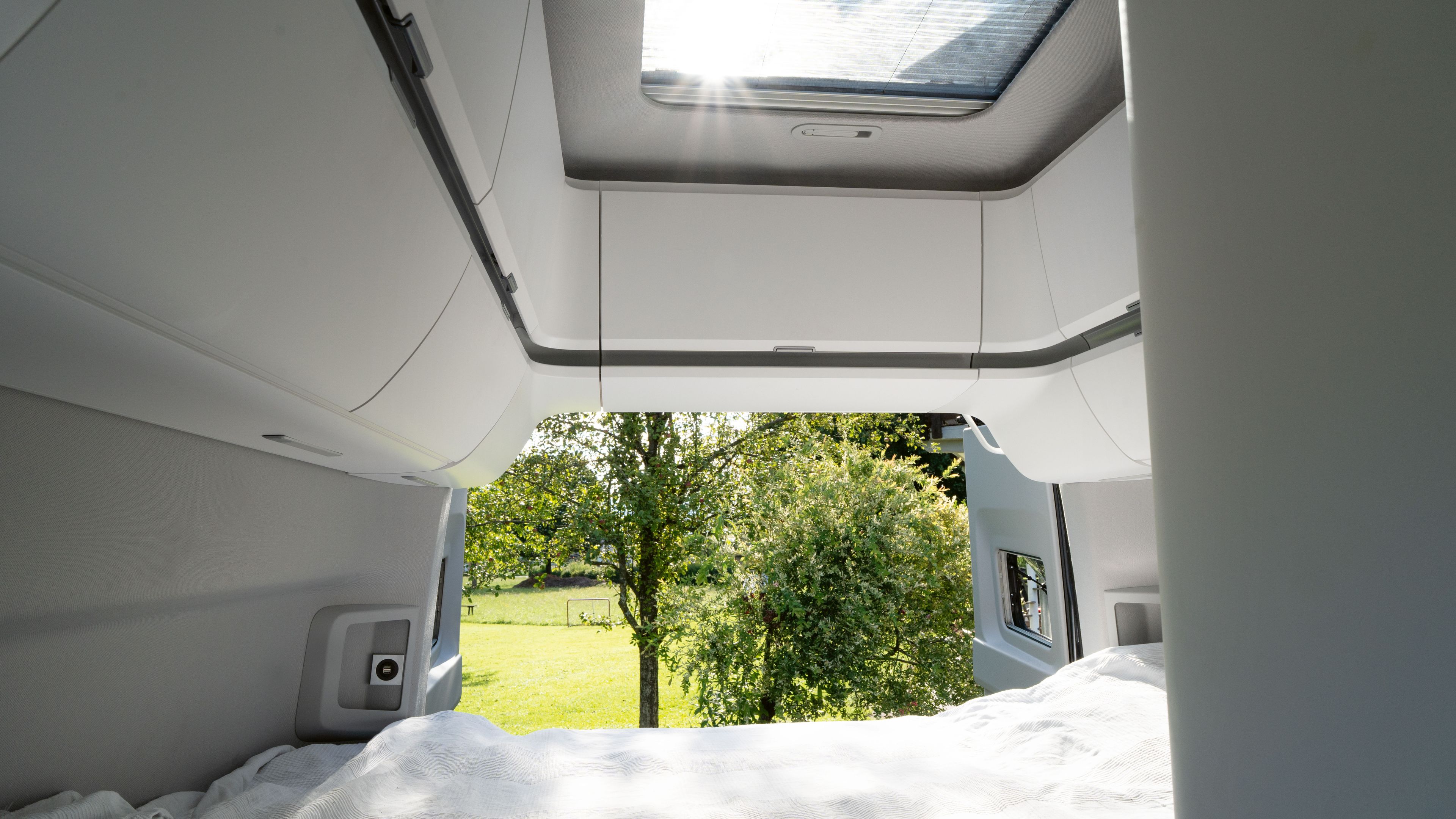 Blick aus einem Wohnmobil mit einem Bett im Vordergrund und grünem Rasen und Bäumen draußen. Das Dachfenster des Fahrzeugs lässt helles Sonnenlicht herein. Links und rechts sind weiße Schränke zu erkennen.