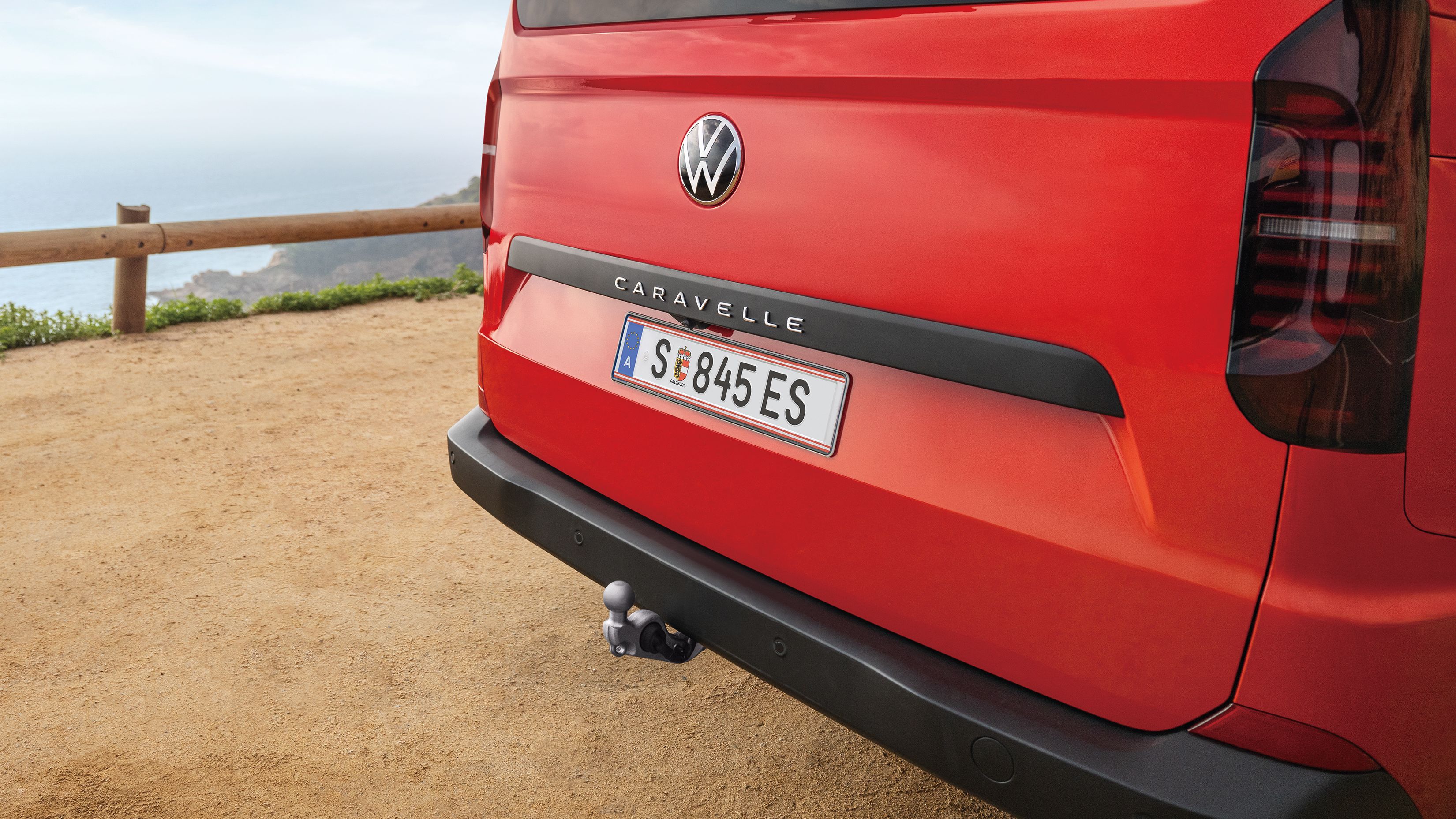 Задната част на червен Volkswagen Caravelle с теглич, спрян на живописен път с изглед към морето