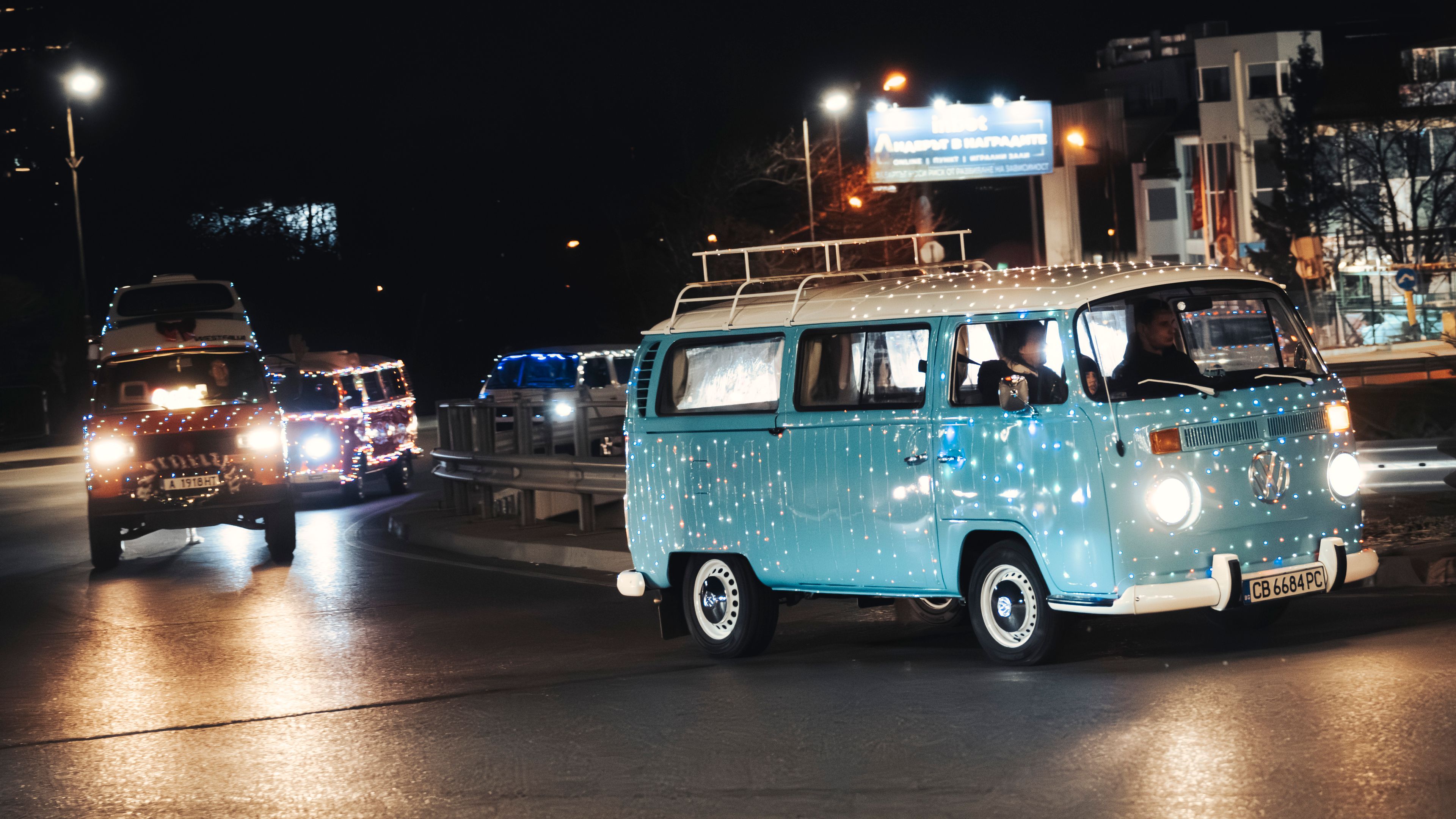 На изображението се виждат два Volkswagen микробуса, украсени със светлини, движещи се по градска улица през нощта. Сградите и рекламен билборд са видими на заден план. Атмосферата е празнична и оживена.