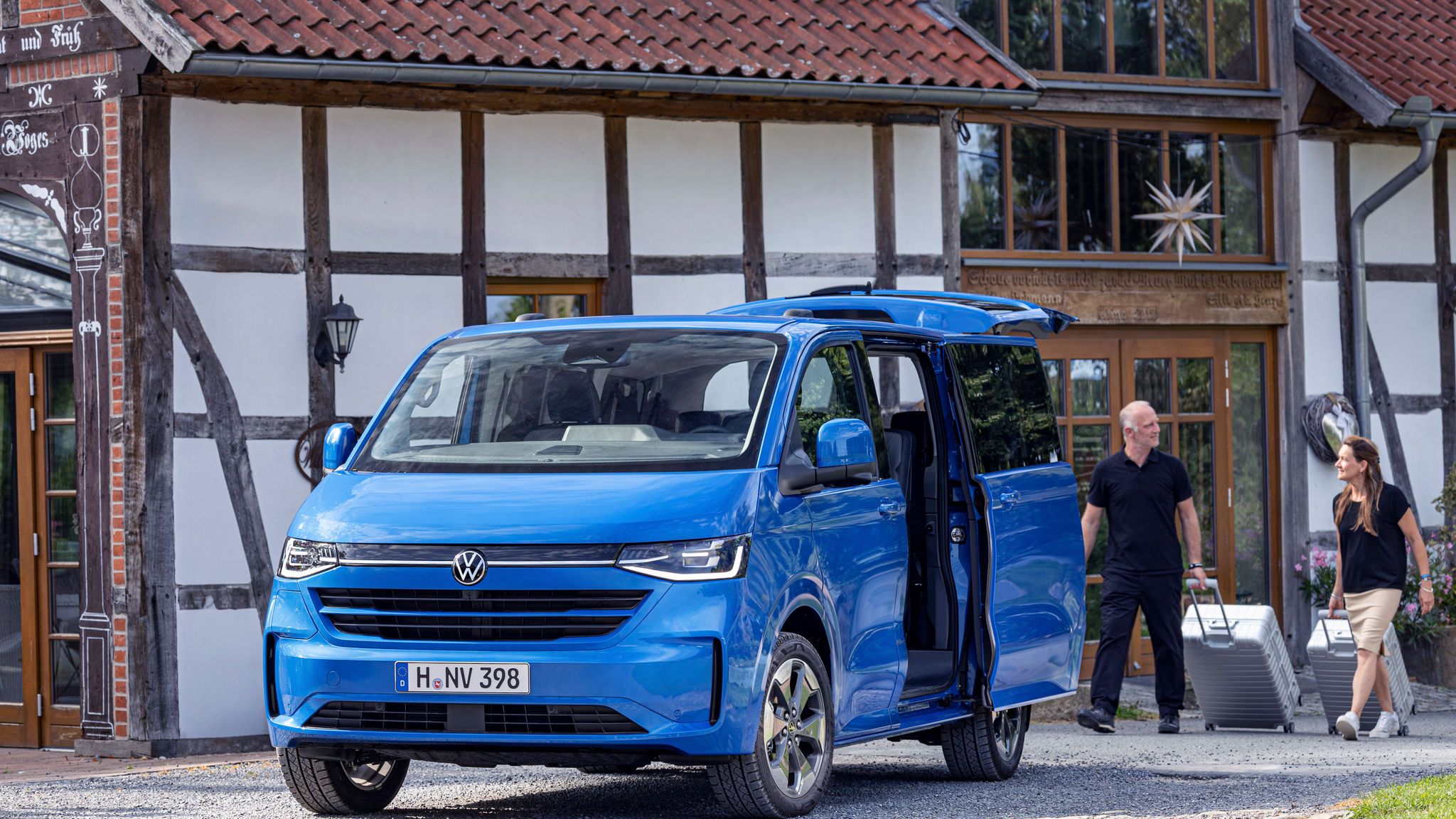 Син Volkswagen Multivan паркиран пред традиционна сграда, с хора, които товарят багаж.