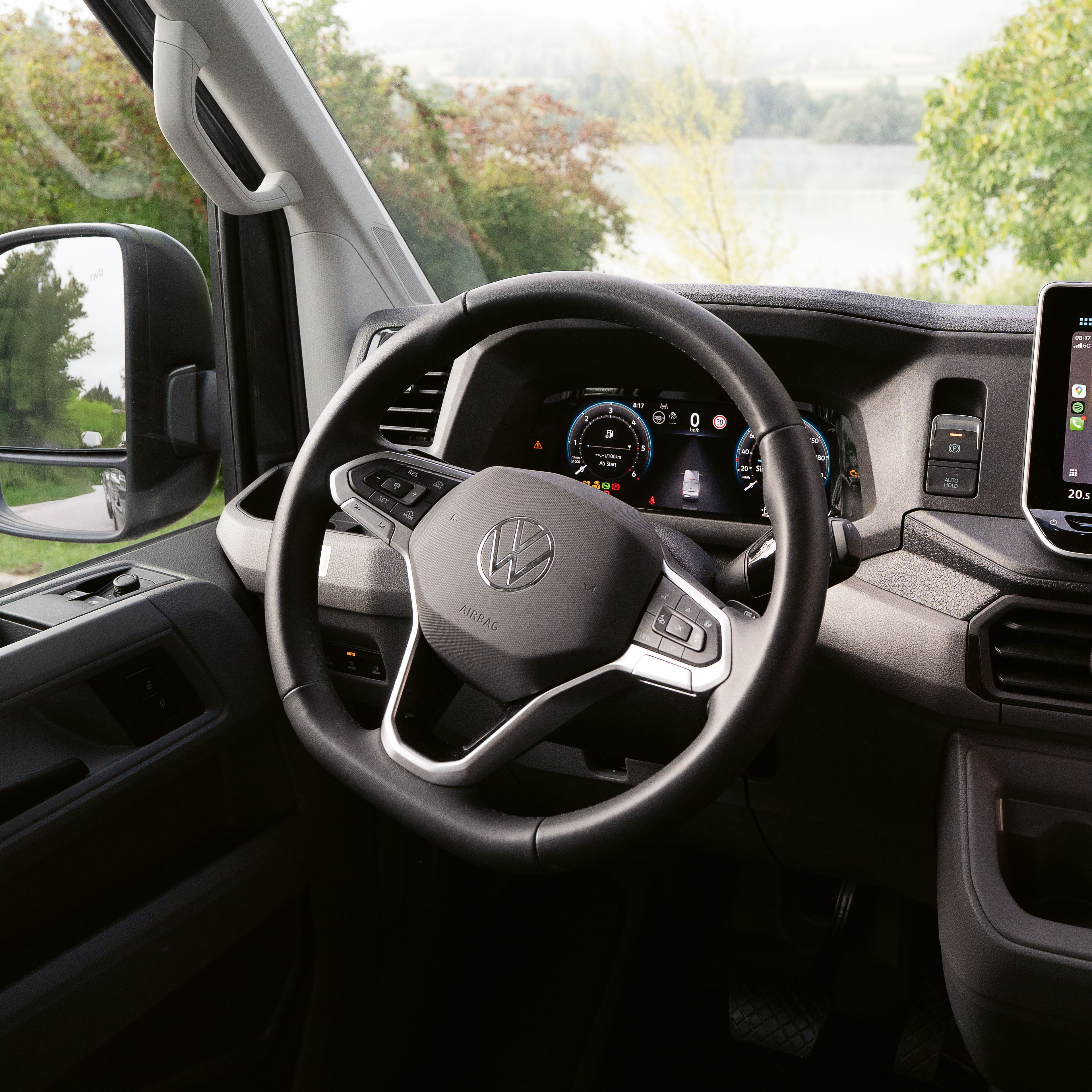 Innenraum eines modernen Autos, das auf einen See blickt. Im Mittelpunkt steht das Lenkrad und ein großes Touchscreen-Display auf dem Armaturenbrett. Durch die Fenster ist eine grüne Landschaft mit Bäumen und Wasser zu sehen.