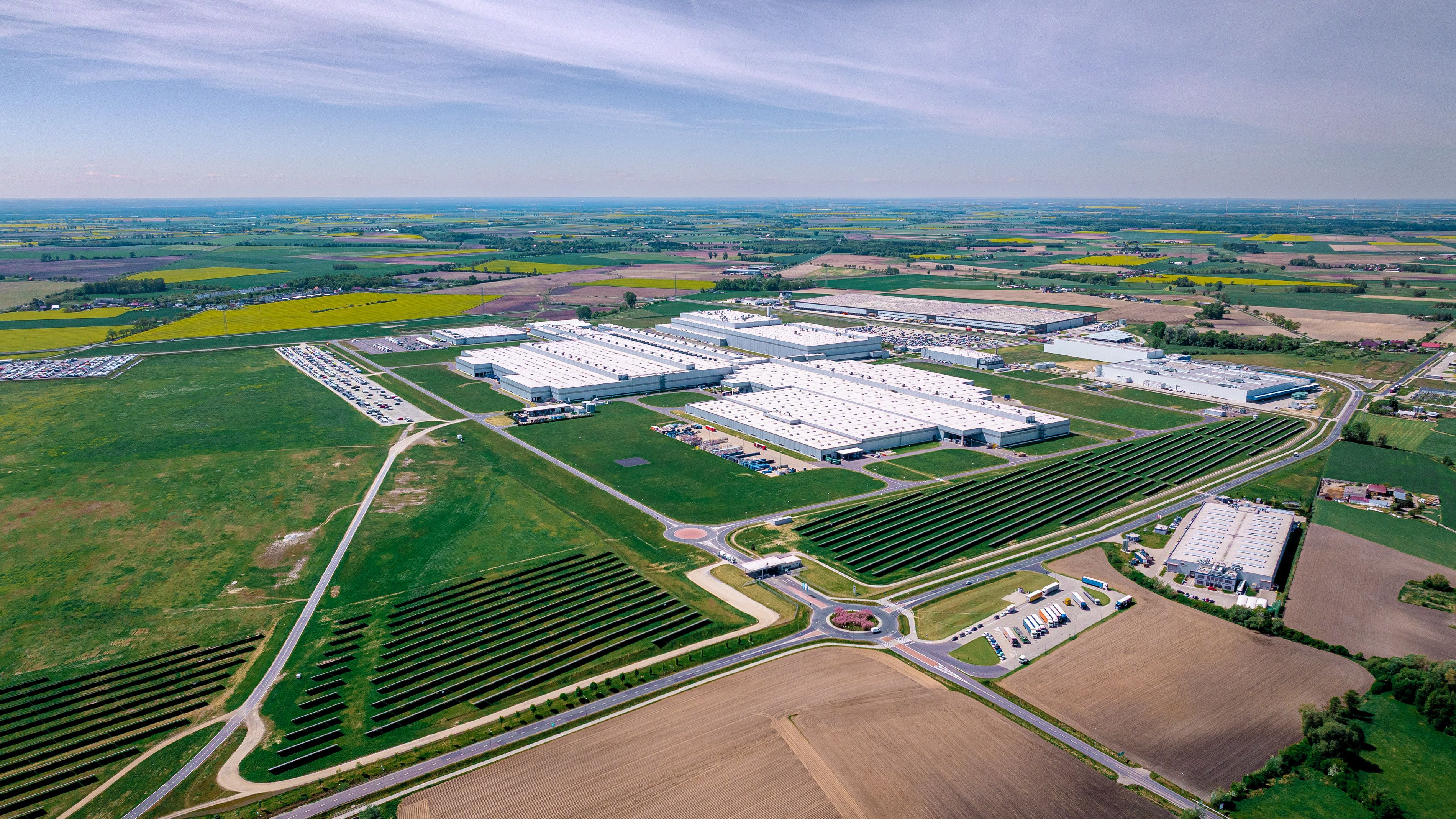 Въздушна снимка на промишлен комплекс, разположен сред зелени и жълти полета в селски район (Вжешня). Комплексът включва няколко големи сгради с бели покриви и добре поддържани зелени площи около тях. В близост се виждат селскостопански полета и пътища, свързващи различни участъци на комплекса.