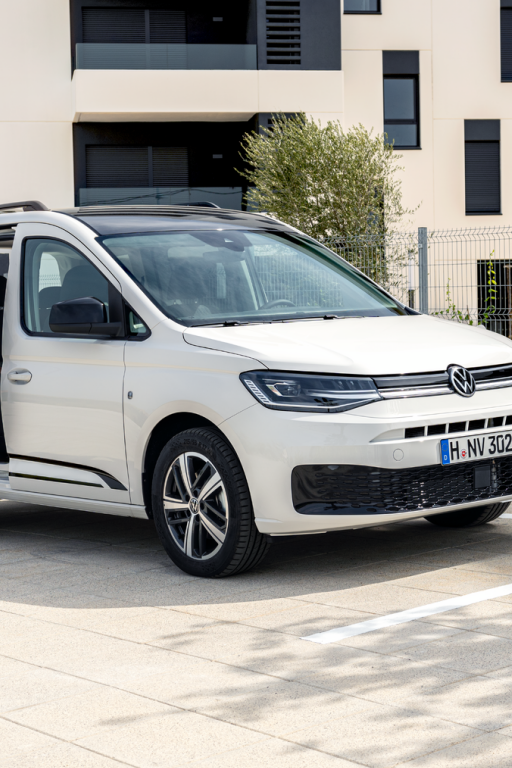 Семейство с детска количка зарежда бял електрически Volkswagen Caddy на зарядна станция пред жилищна сграда