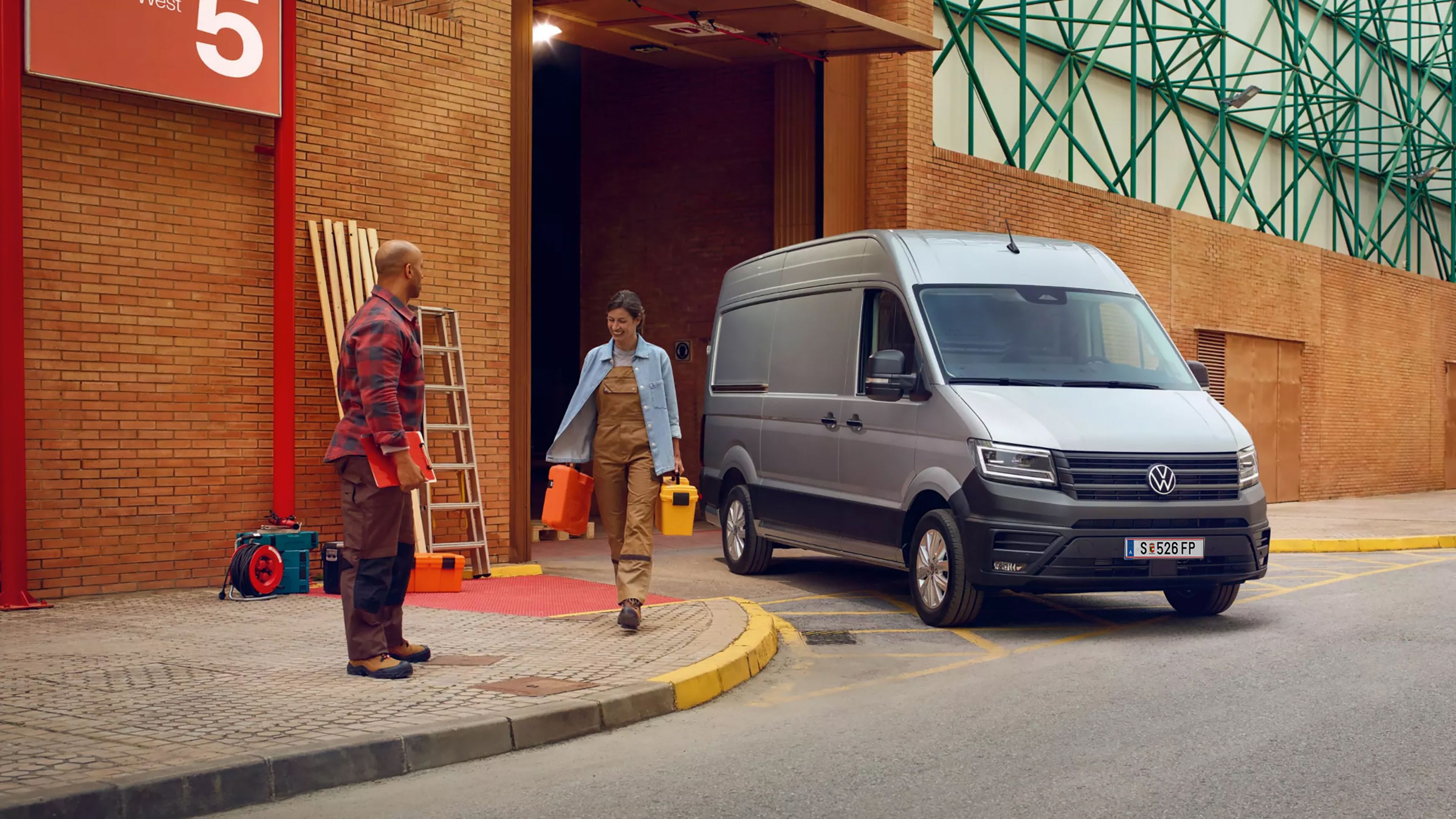 Der neue Crafter Двама работници пред сграда със сив Volkswagen Crafter, паркиран на товарна рампа.