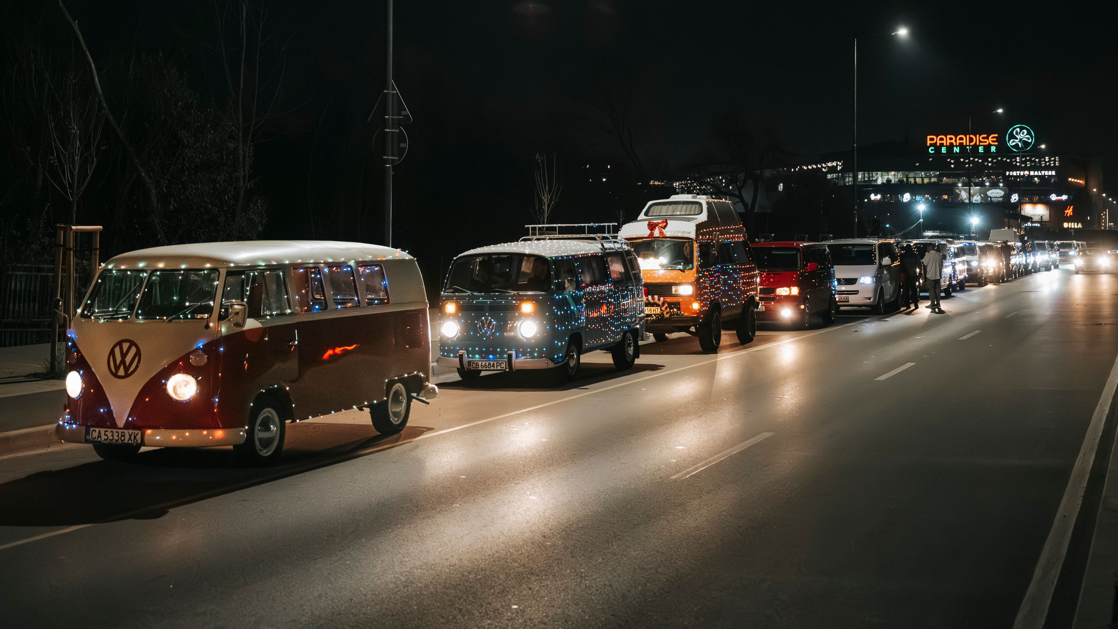 Нощна сцена на колона от бусове Volkswagen, украсени със светлини по стъкларията. Бусовете се движат по осветен градски път. В далечината се вижда светещ знак на мол “Paradise Center”.