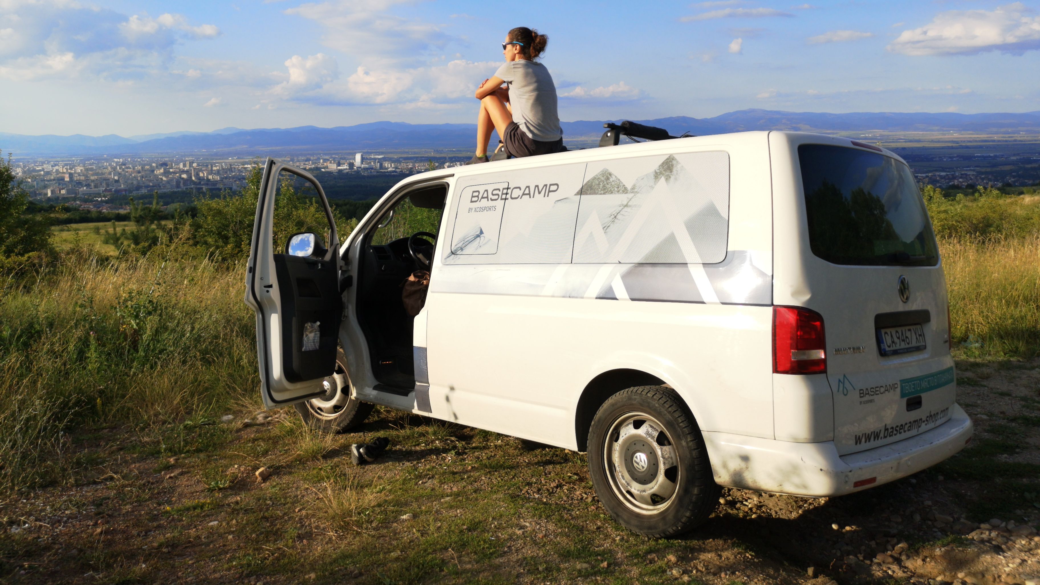 Жена седи върху покрива на бял ван с отворена предна врата, наслаждавайки се на красивата гледка. В далечината се вижда град с множество сгради, заобиколен от хълмове и облачно небе.