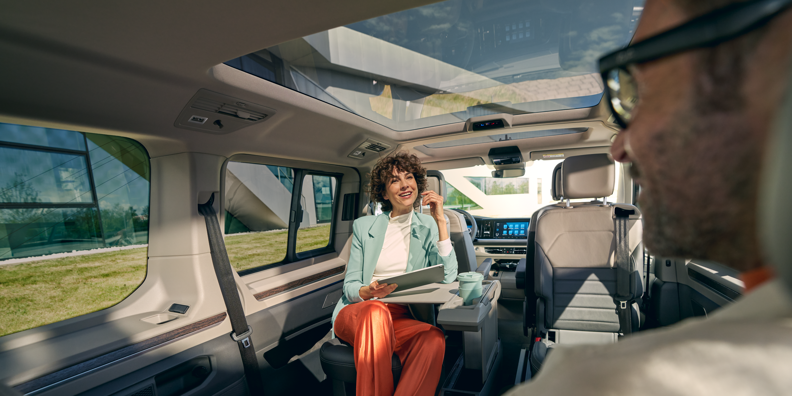 Просторен и модерен интериор на Volkswagen Multivan Жена с таблет разговаря с мъж в просторния интериор на Volkswagen Multivan с панорамен покрив.
