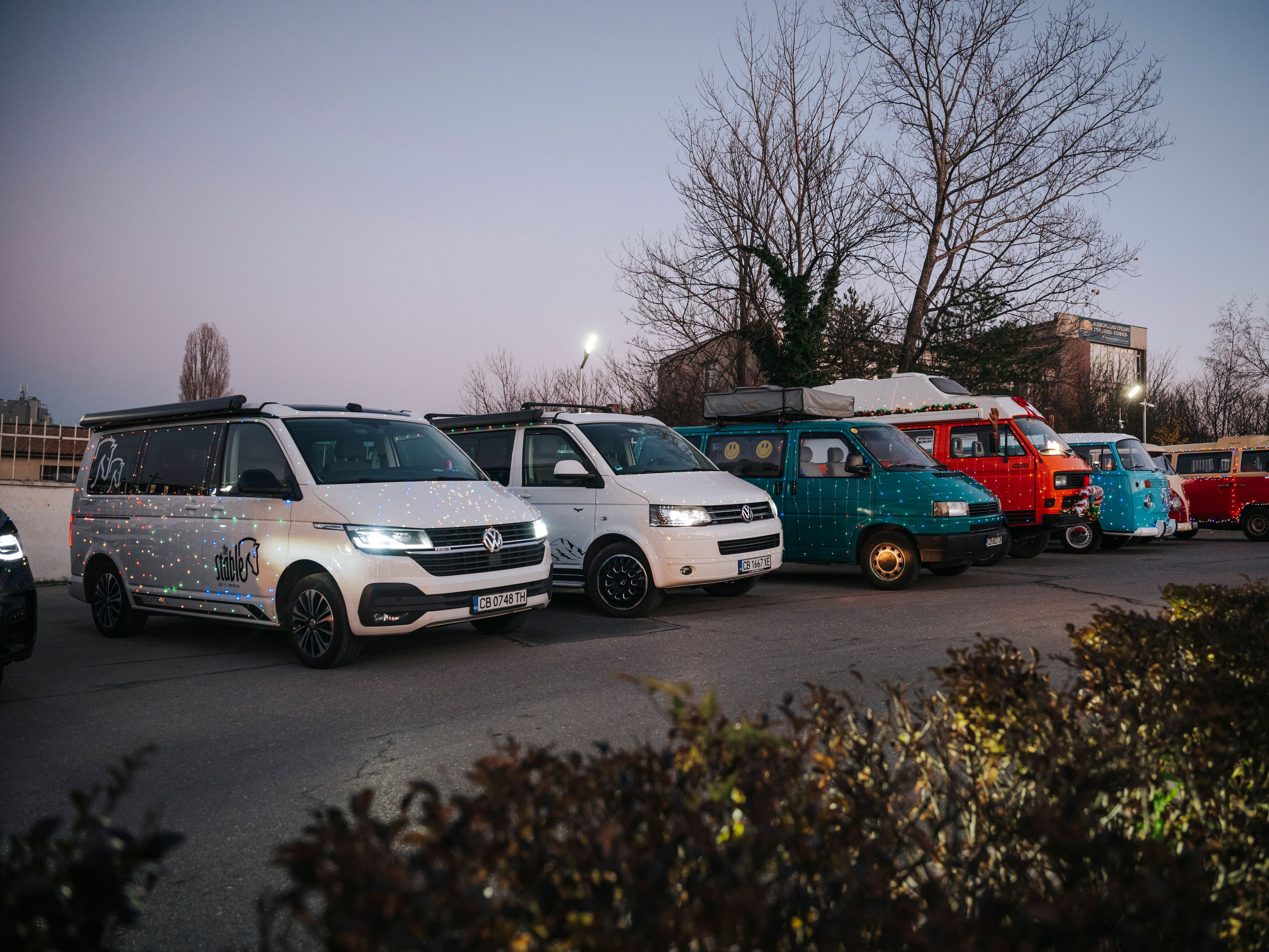 Редица кемпери и микробуси Volkswagen са паркирани на открит паркинг по здрач. На преден план има растения, а отзад са видими голи дървета и сграда. Някои от превозните средства са ярко оцветени и имат различни дизайни.