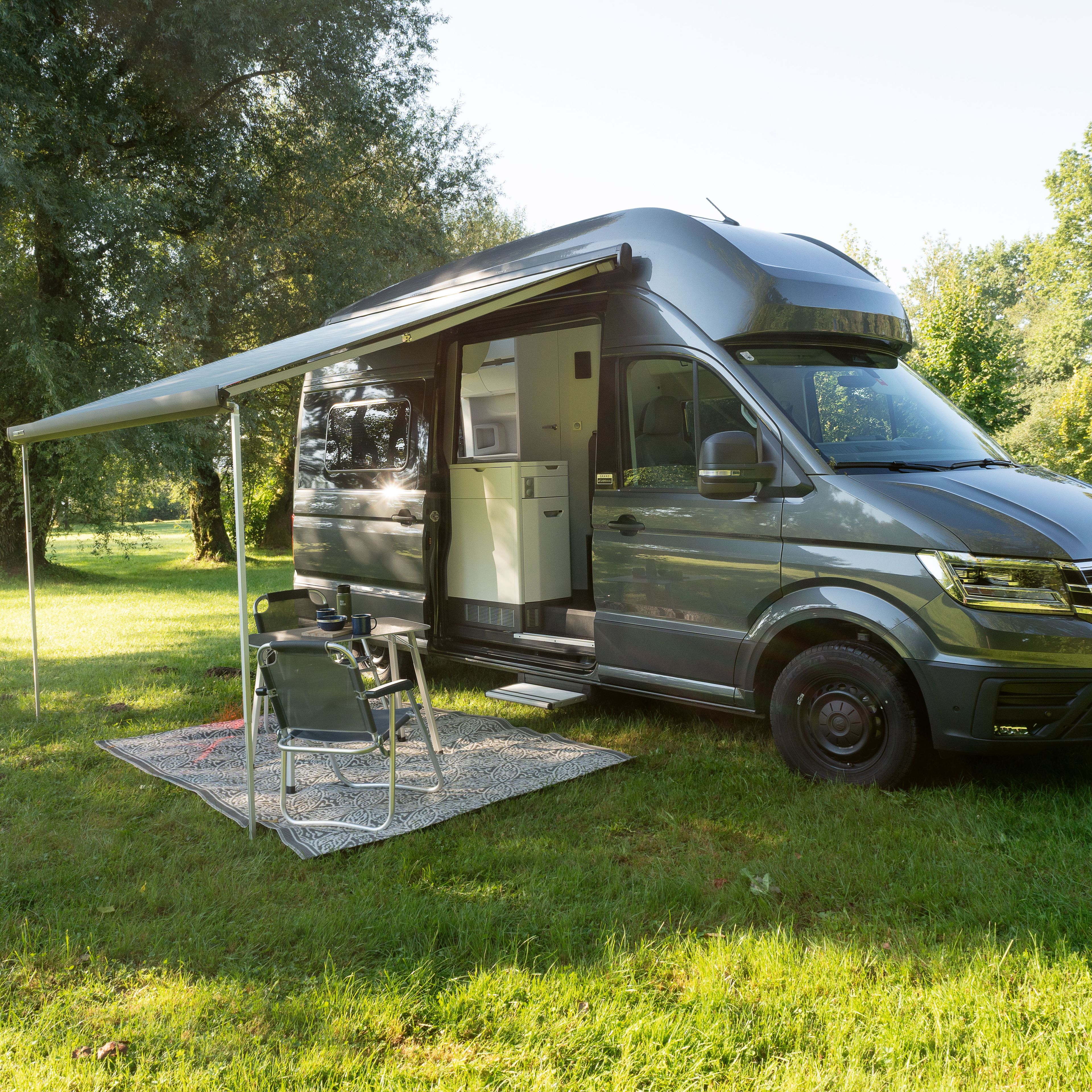 Ein großer grauer Wohnmobil steht auf einer grünen Wiese mit Bäumen im Hintergrund. Das Fahrzeug hat eine ausgefahrene Markise, unter der ein Tisch und zwei Stühle stehen. Im Hintergrund ist eine für Camping geeignete Naturumgebung.