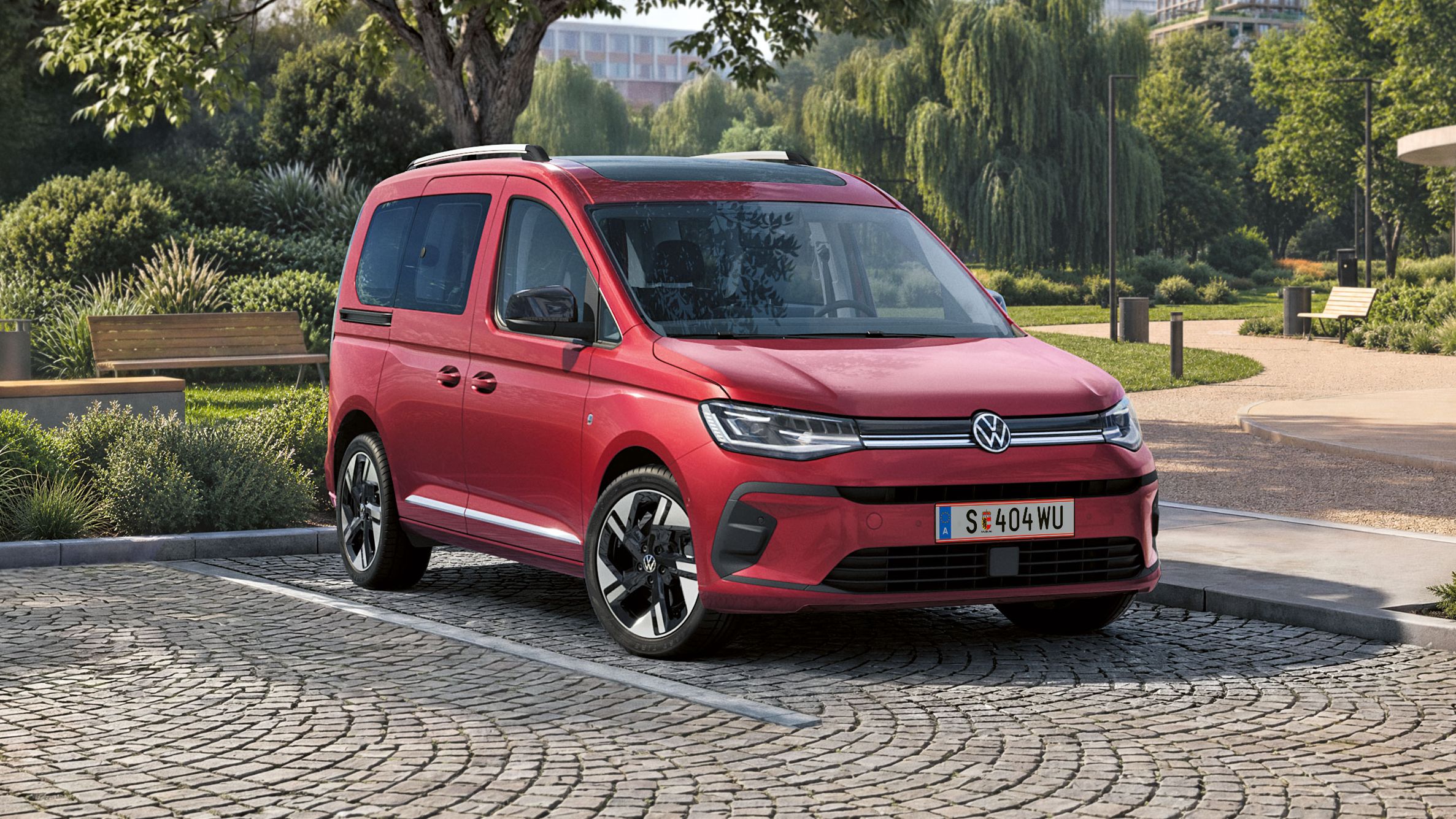 Червен миниван Volkswagen е паркиран на павиран паркинг в модерна градска градина с много зеленина и дървета. На заден план се виждат високи офис сгради и алеи с добре поддържан ландшафтен дизайн. Сцената е при дневна светлина и подчертава семейния и градски характер на автомобила.