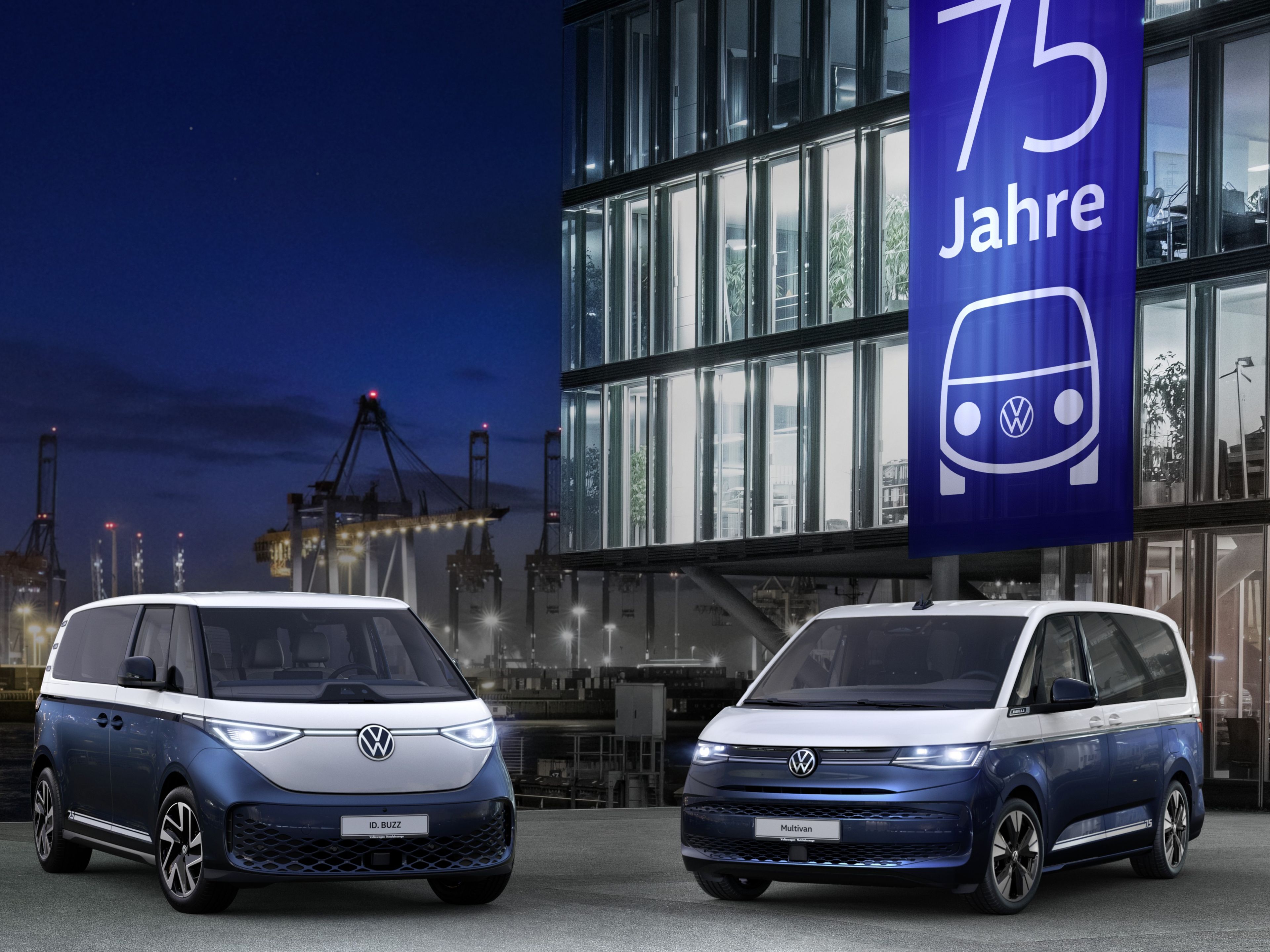 На изображението са показани два сини модела ID.Buzz и Multivan, паркирани пред модерна сграда през нощта. Сградата е осветена отвътре и отвън и има голям банер с надпис "75 Jahre", изобразяващ логото на VW. На заден план се вижда пристанище с кранове и осветени кораби.
