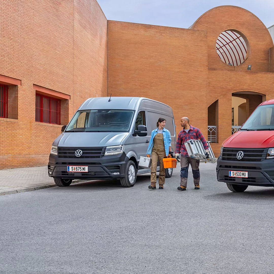 Два товарни автомобила Volkswagen Crafter – един закрит сив ван и един червен с открита платформа, паркирани на строителен обект с двама работници