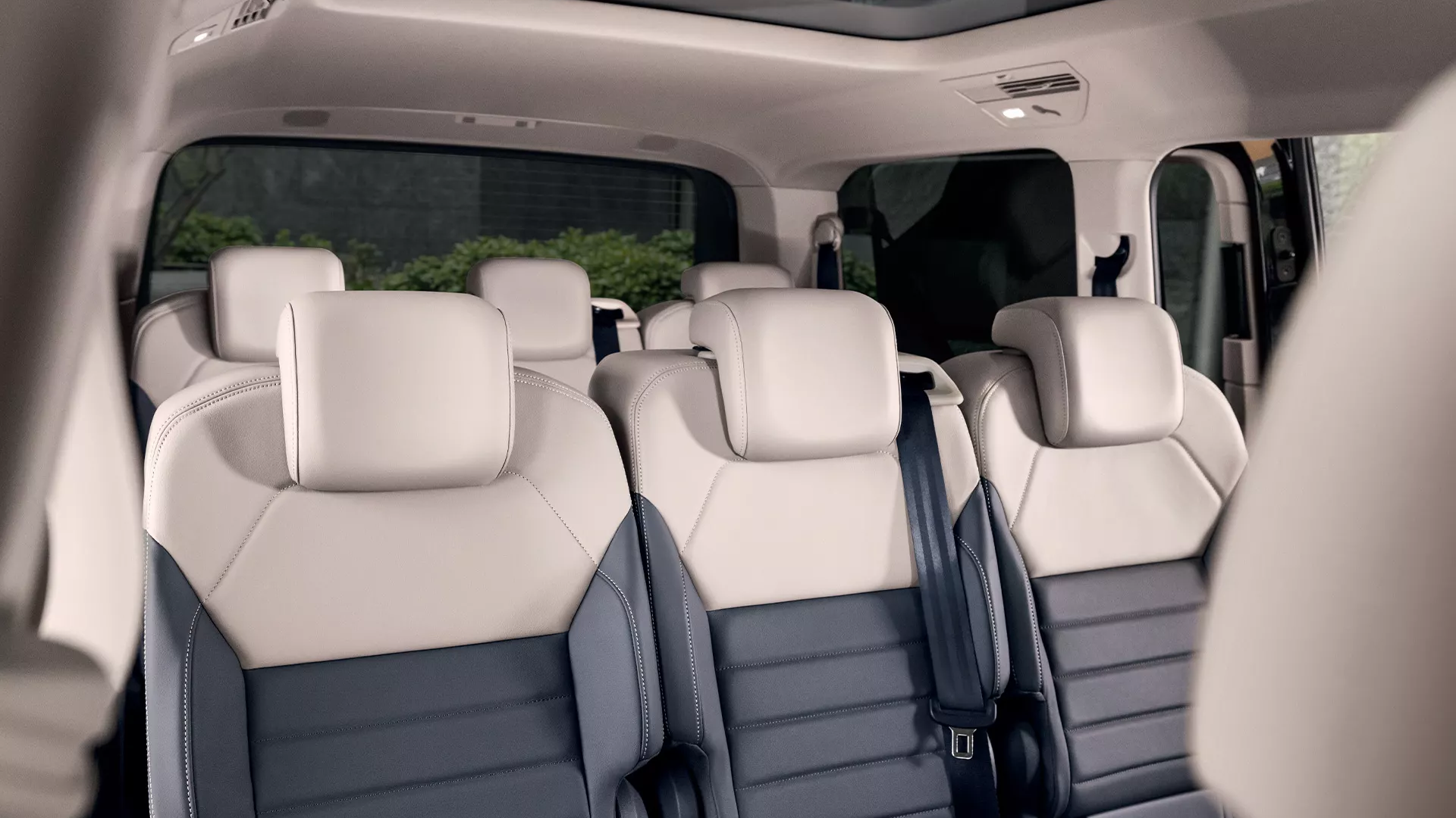 Innenraum eines Multivans mit Ledersitzen. Die Sitze sind beige und dunkelgrau, und das Fahrzeug hat ein Panoramadach. Die Rückbank hat Platz für drei Personen.