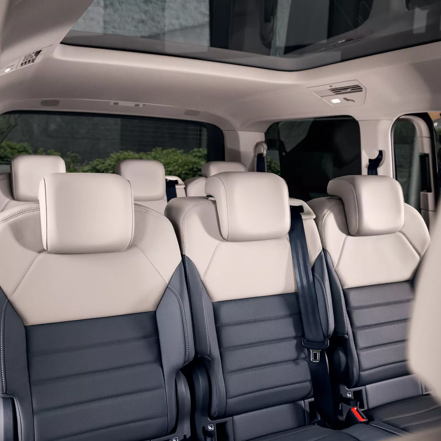 Innenraum eines Multivans mit Ledersitzen. Die Sitze sind beige und dunkelgrau, und das Fahrzeug hat ein Panoramadach. Die Rückbank hat Platz für drei Personen.