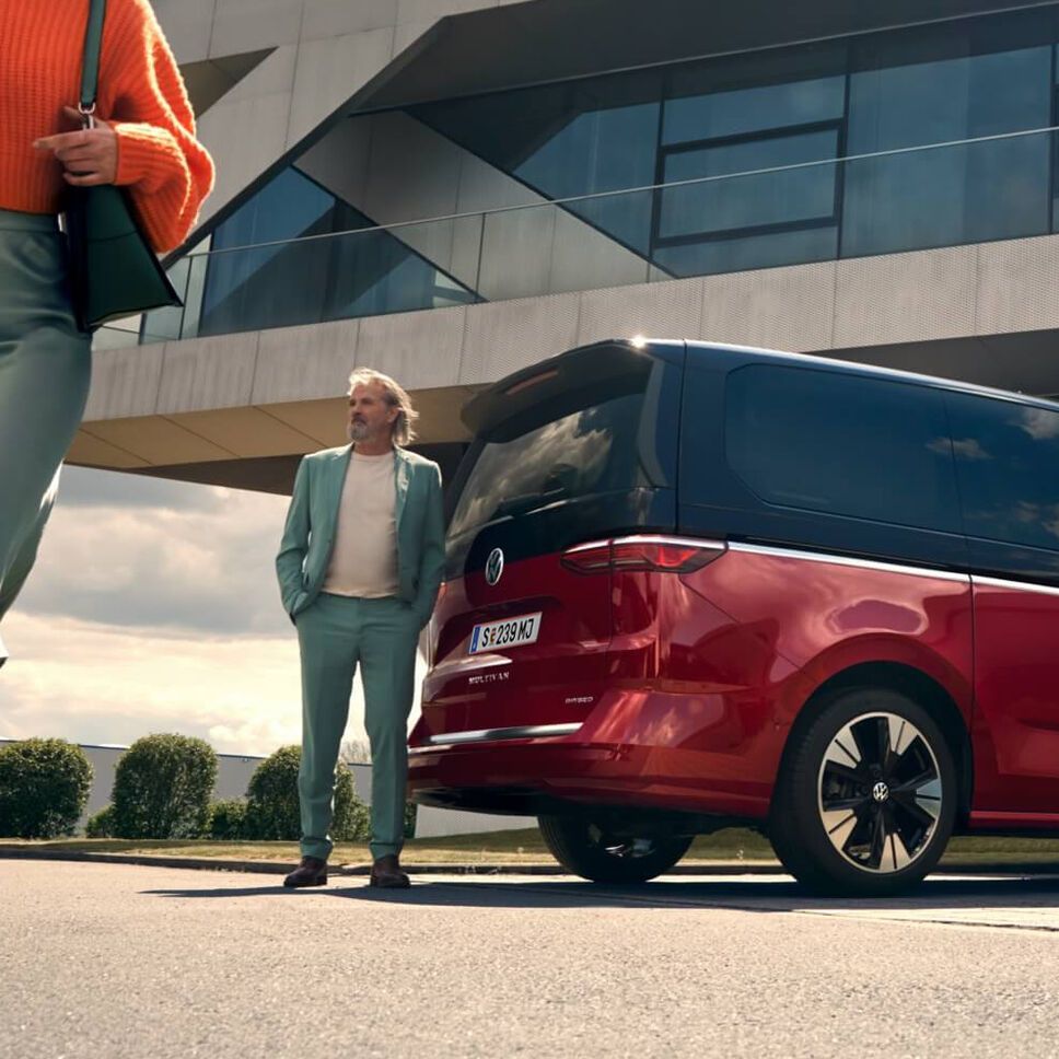 Червен Volkswagen Multivan, свързан към електрическа зарядна станция пред модерна сграда.