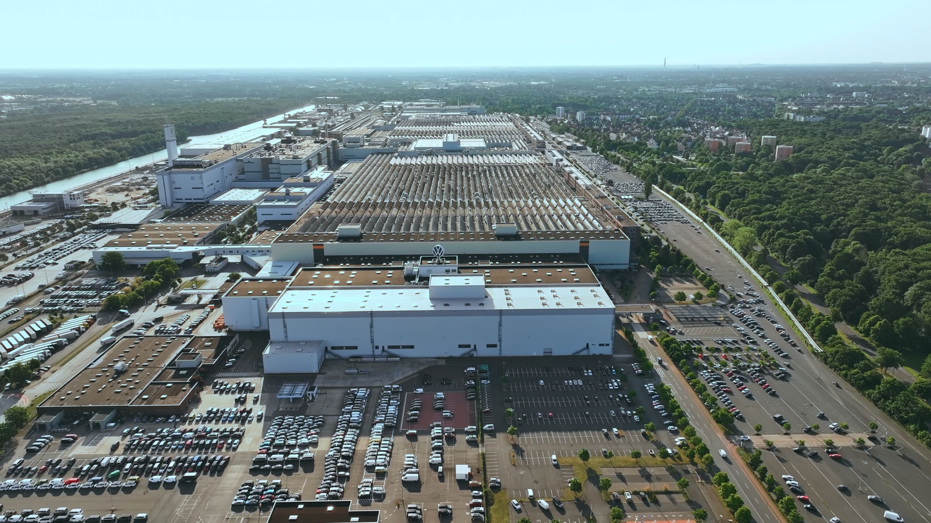 Въздушен изглед към голям индустриален Volkswagen завод с множество фабрични сгради, паркирани автомобили пред тях и зелена зона в далечината.