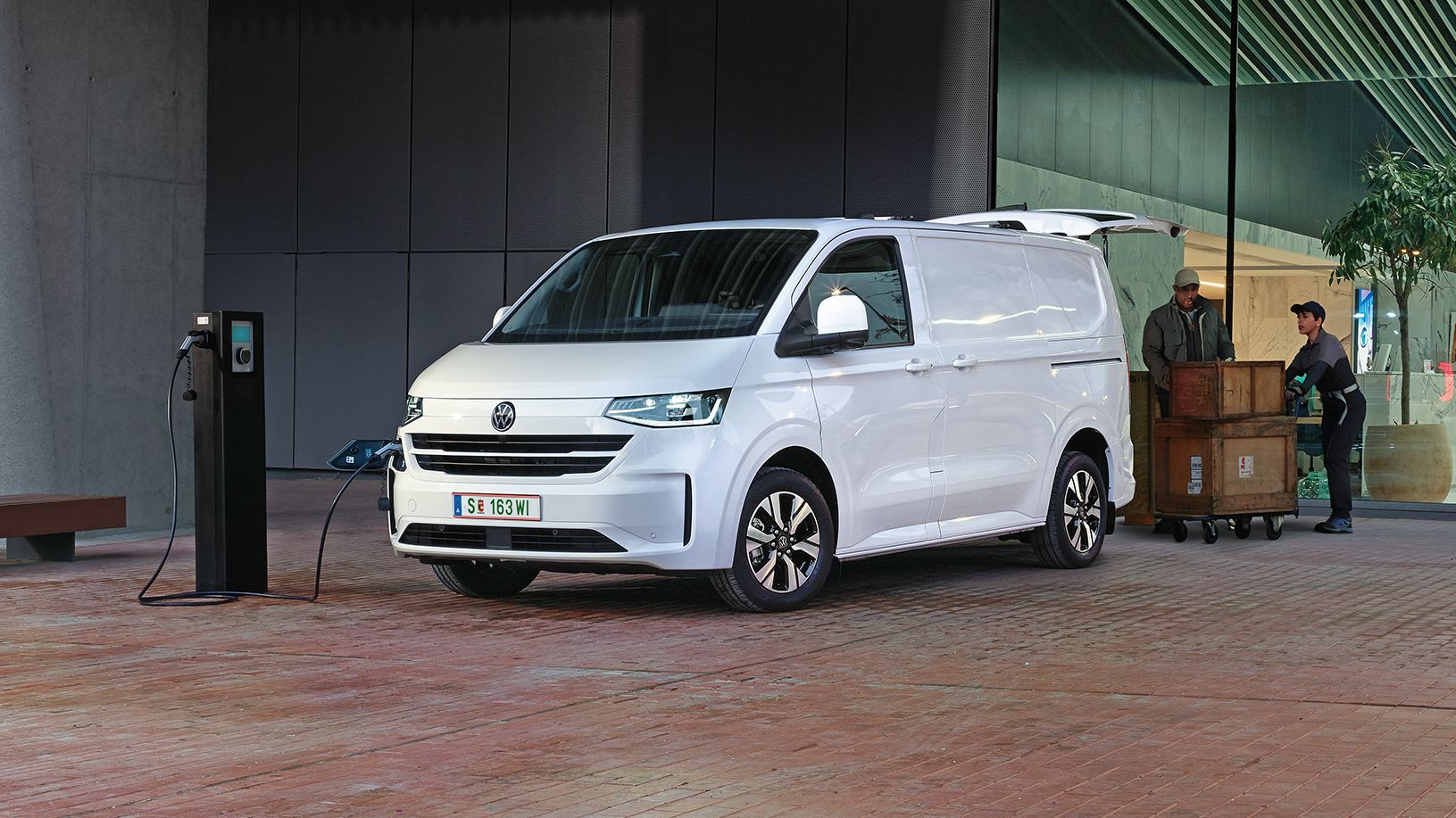 Електрически Volkswagen Transporter, включен към зарядна станция пред офис сграда, докато се разтоварват стоки.