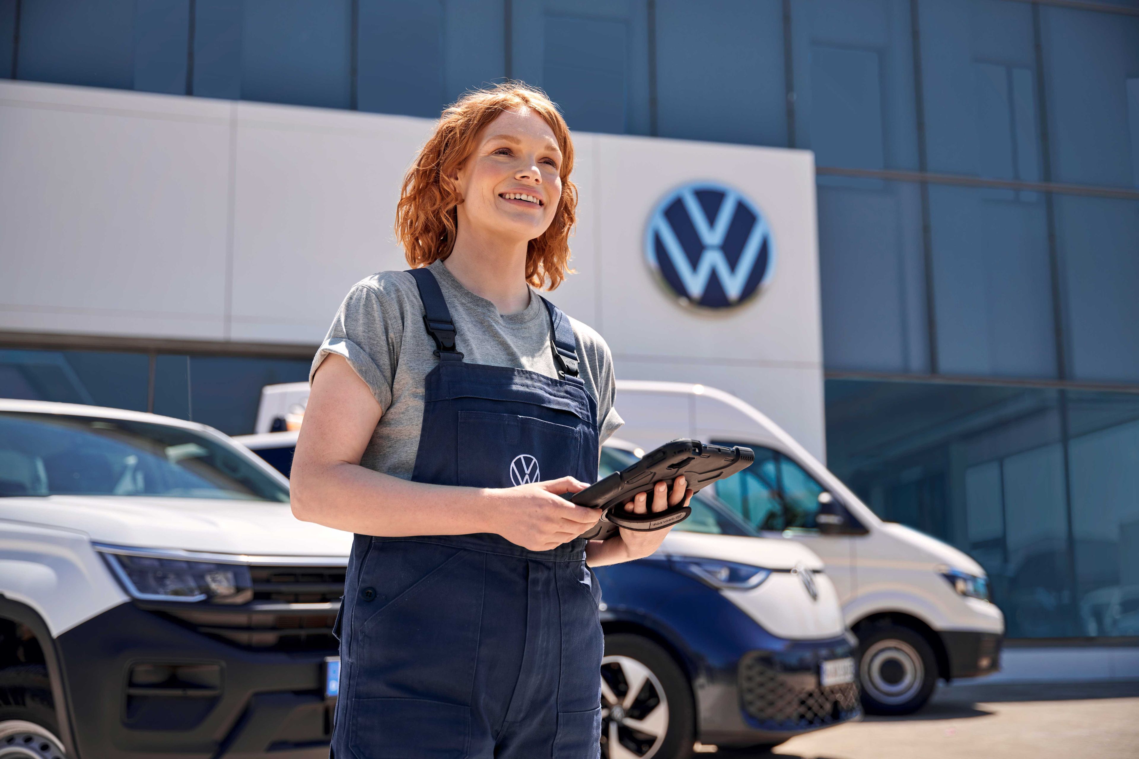 Жена, облечена в работен гащеризон, държи таблет пред сграда с логото на Volkswagen. Зад нея има паркирани автомобил и бус на Volkswagen.