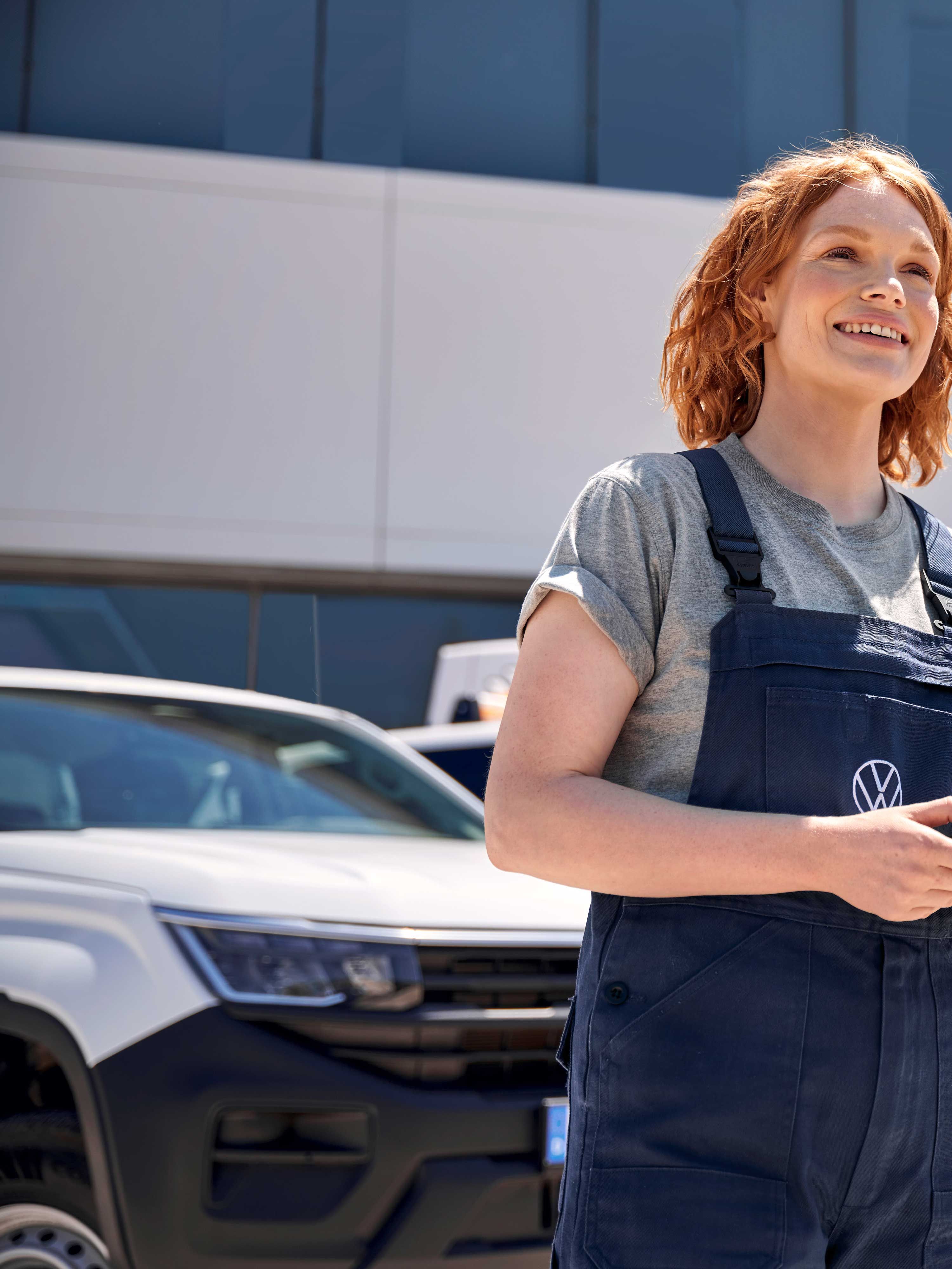 Жена, облечена в работен гащеризон, държи таблет пред сграда с логото на Volkswagen. Зад нея има паркирани автомобил и бус на Volkswagen.