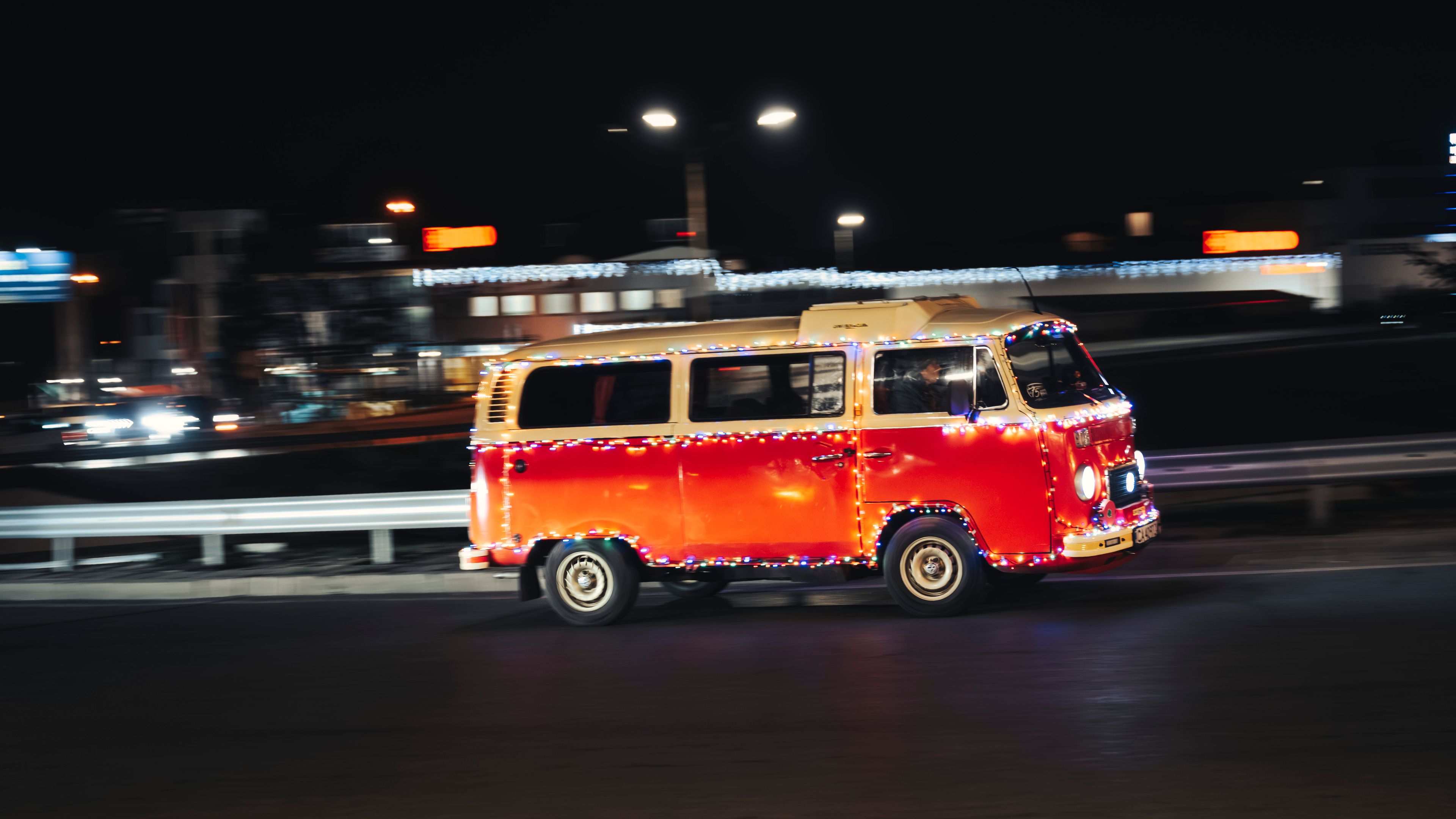 Оранжев Volkswagen микробус, украсен с цветни светлини, се движи по градска улица през нощта. Фоновите светлини на града са размазани поради движението на микробуса.