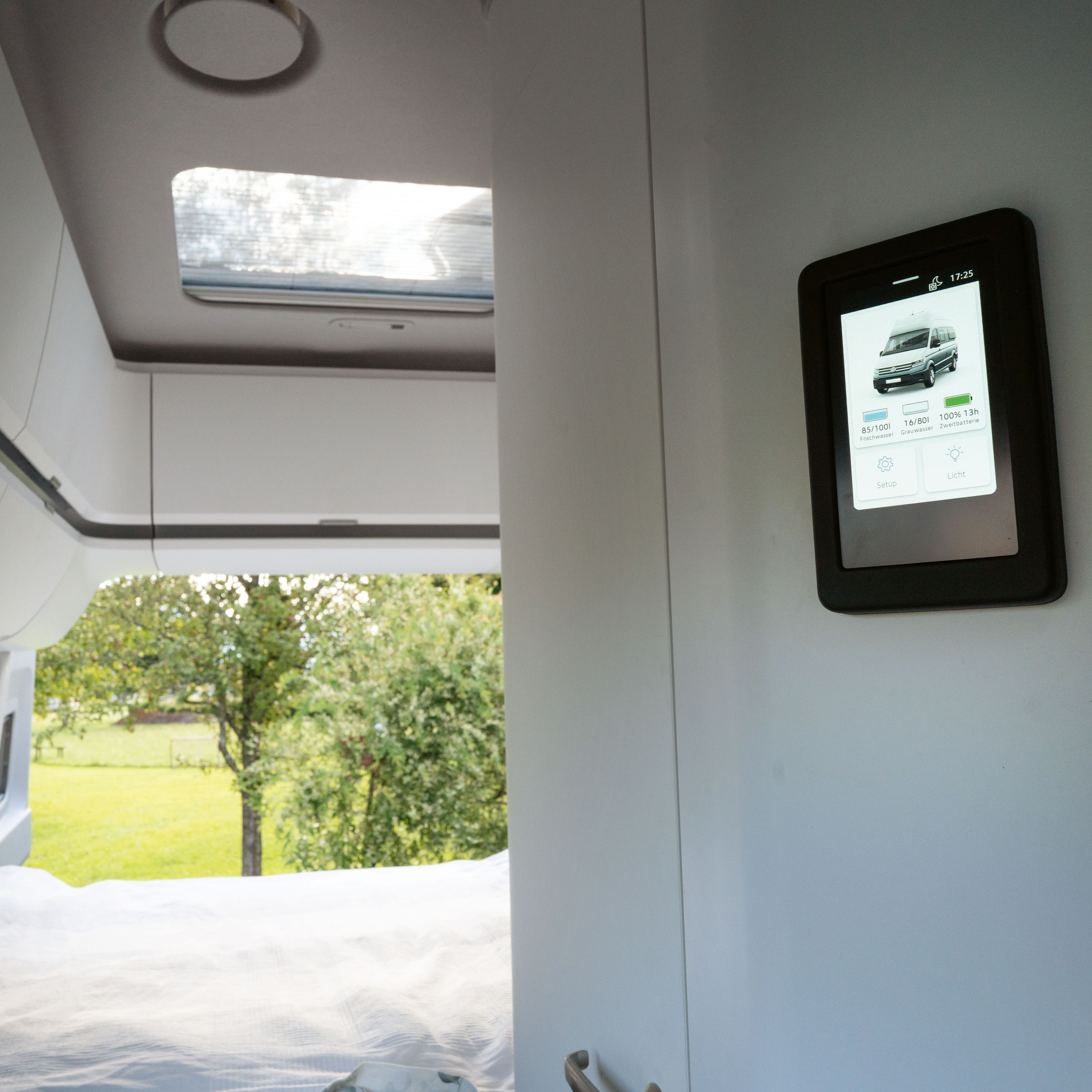 Blick in das Innere eines Wohnmobils mit einem Bett und einem digitalen Bildschirm. Der Bildschirm zeigt ein Bild eines Autos und einige Informationen. Im Hintergrund ist durch ein Fenster eine grüne Landschaft sichtbar.