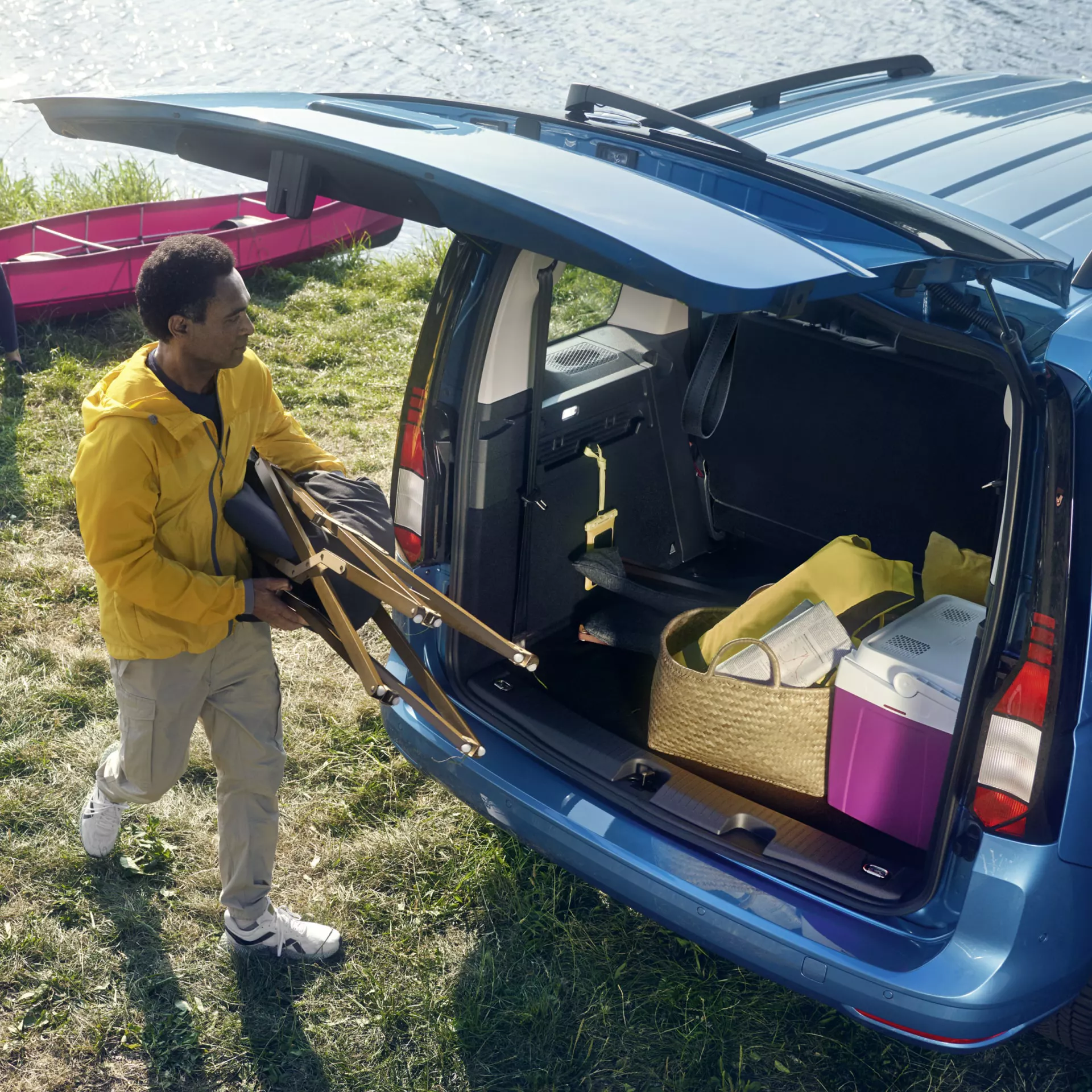 Мъж в яркожълто яке, носещ столове, приближава син автомобил с отворен багажник, паркиран близо до воден съд. В багажника има туристически принадлежности като кутия за храна и кошница. На заден план също се вижда розова лодка.
