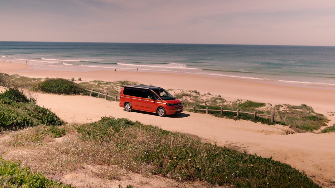 Червен Volkswagen Multivan на пясъчен път край морето, с гледка към плажа и вълните