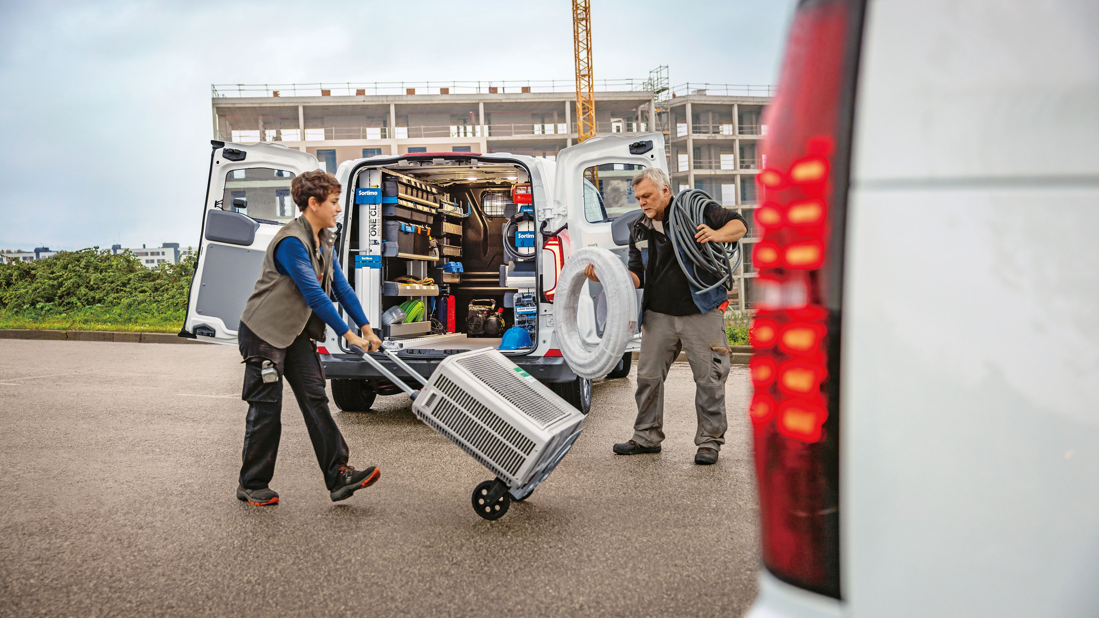 Двама техници разтоварват климатик и оборудване от напълно оборудван Volkswagen Transporter на строителен обект.
