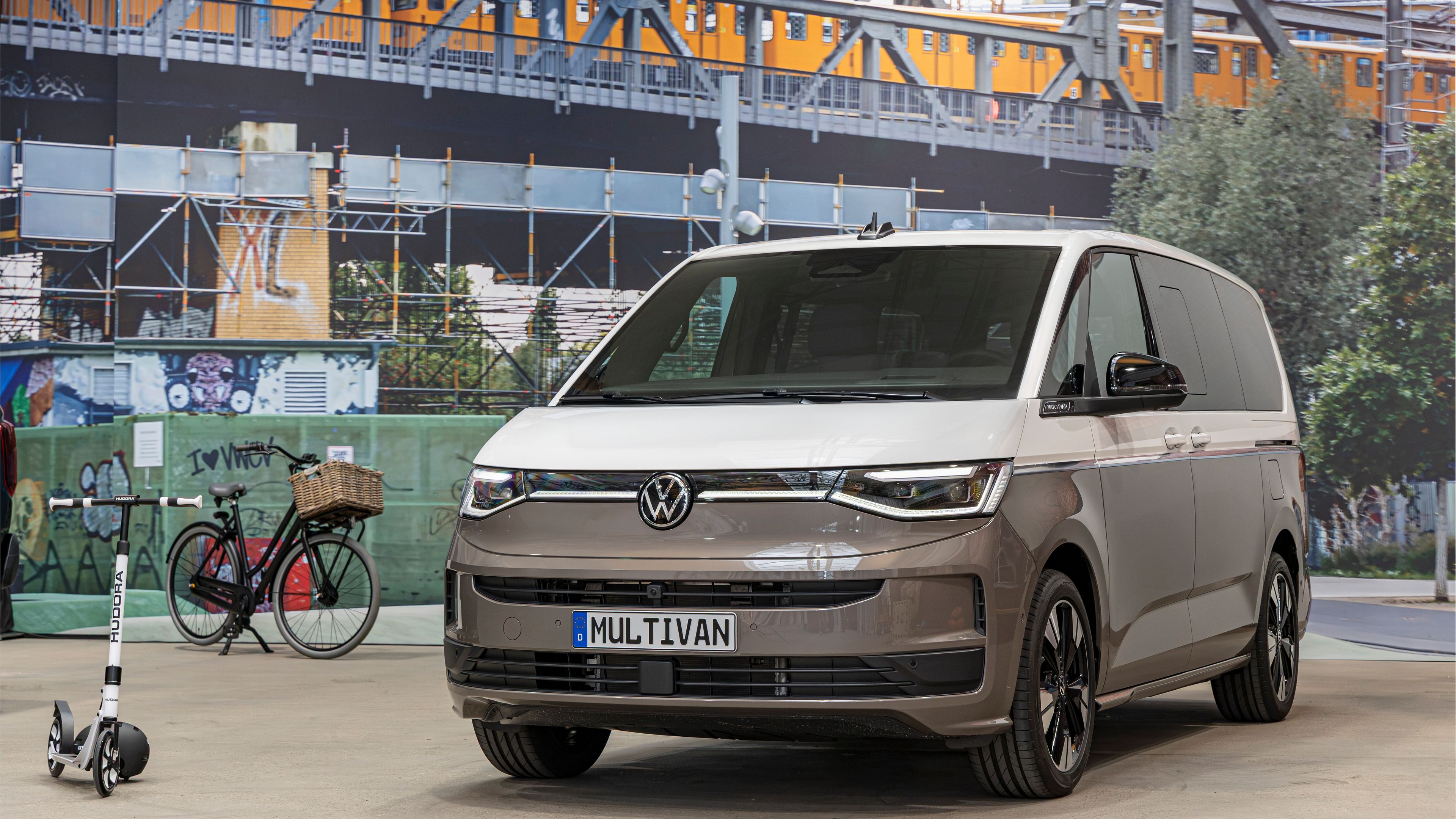 Съвременен миниван Volkswagen Multivan в бежово-сив цвят е паркиран на закрито пред голям стенопис със железопътен мост и преминаващ влак. Вляво има паркирано колело с кошница и тротинетка, оставени до стената. Атмосферата напомня градска среда с акцент върху модерна мобилност.