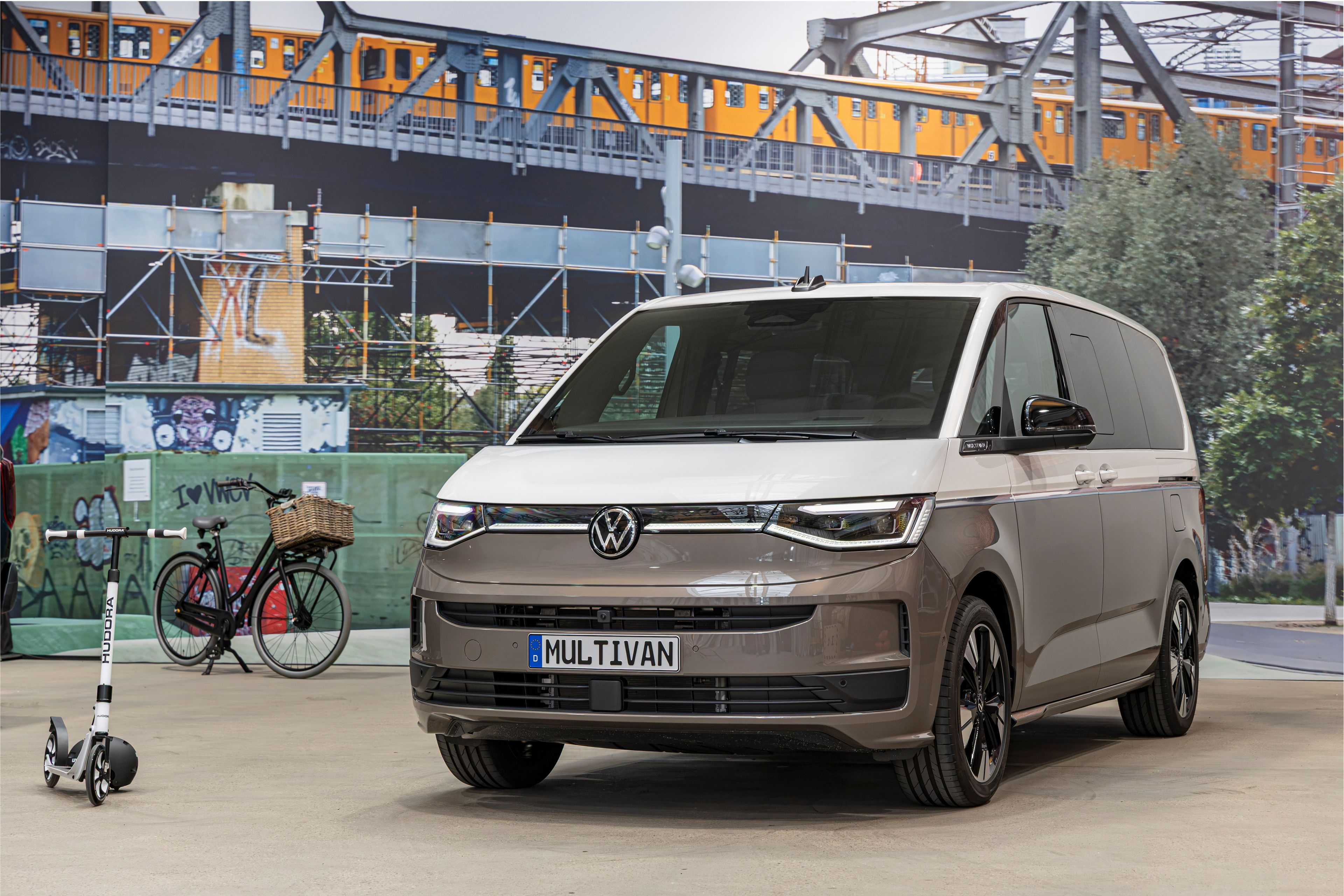 Съвременен миниван Volkswagen Multivan в бежово-сив цвят е паркиран на закрито пред голям стенопис със железопътен мост и преминаващ влак. Вляво има паркирано колело с кошница и тротинетка, оставени до стената. Атмосферата напомня градска среда с акцент върху модерна мобилност.