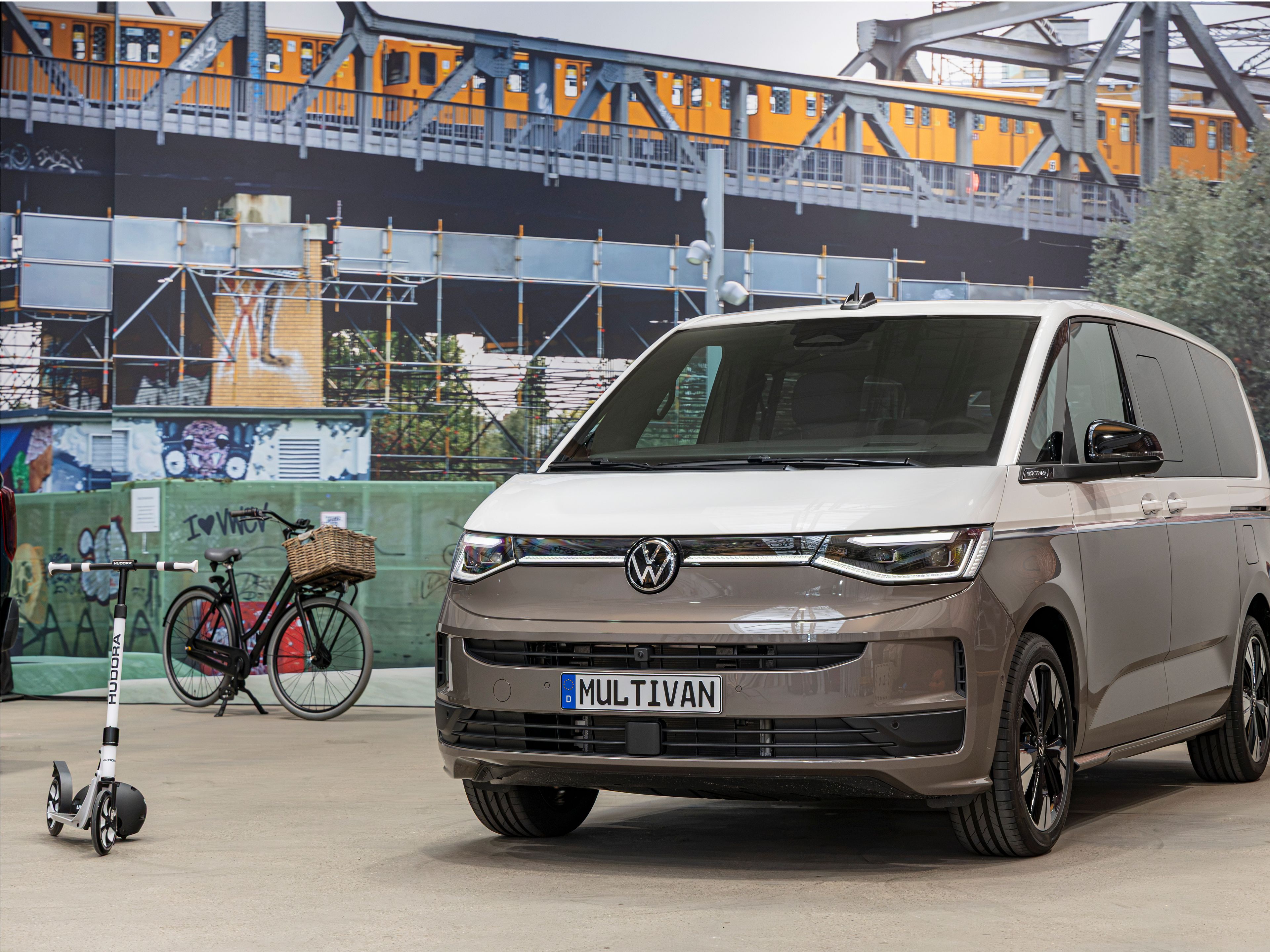 Съвременен миниван Volkswagen Multivan в бежово-сив цвят е паркиран на закрито пред голям стенопис със железопътен мост и преминаващ влак. Вляво има паркирано колело с кошница и тротинетка, оставени до стената. Атмосферата напомня градска среда с акцент върху модерна мобилност.