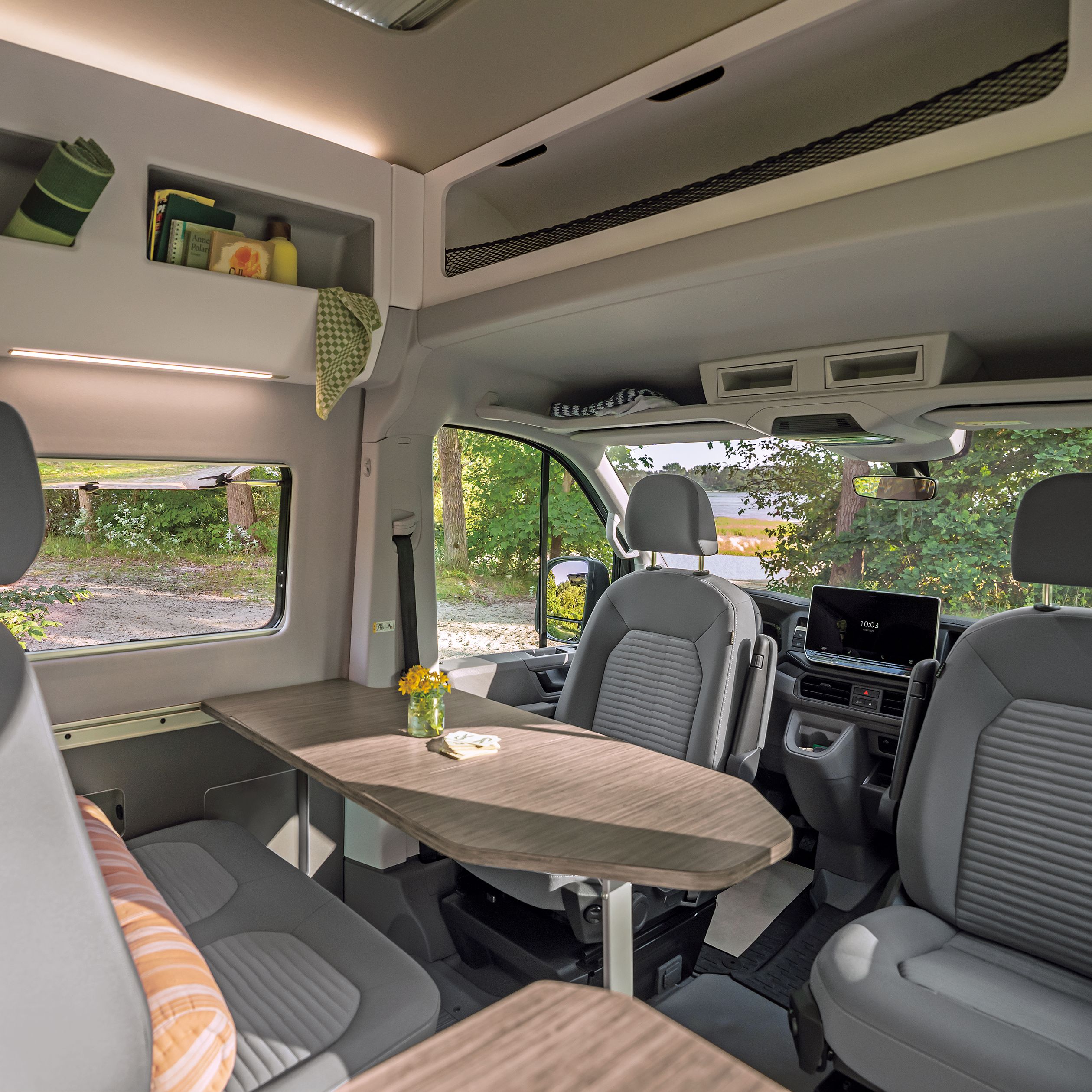 Das Bild zeigt das Innere eines Wohnmobils mit zwei grauen Vordersitzen, einem Tisch und einer kleinen Vase mit Blumen darauf. Im Hintergrund sind Regale mit ein paar Gegenständen zu sehen. Durch das Fenster ist ein grüner Außenbereich sichtbar.