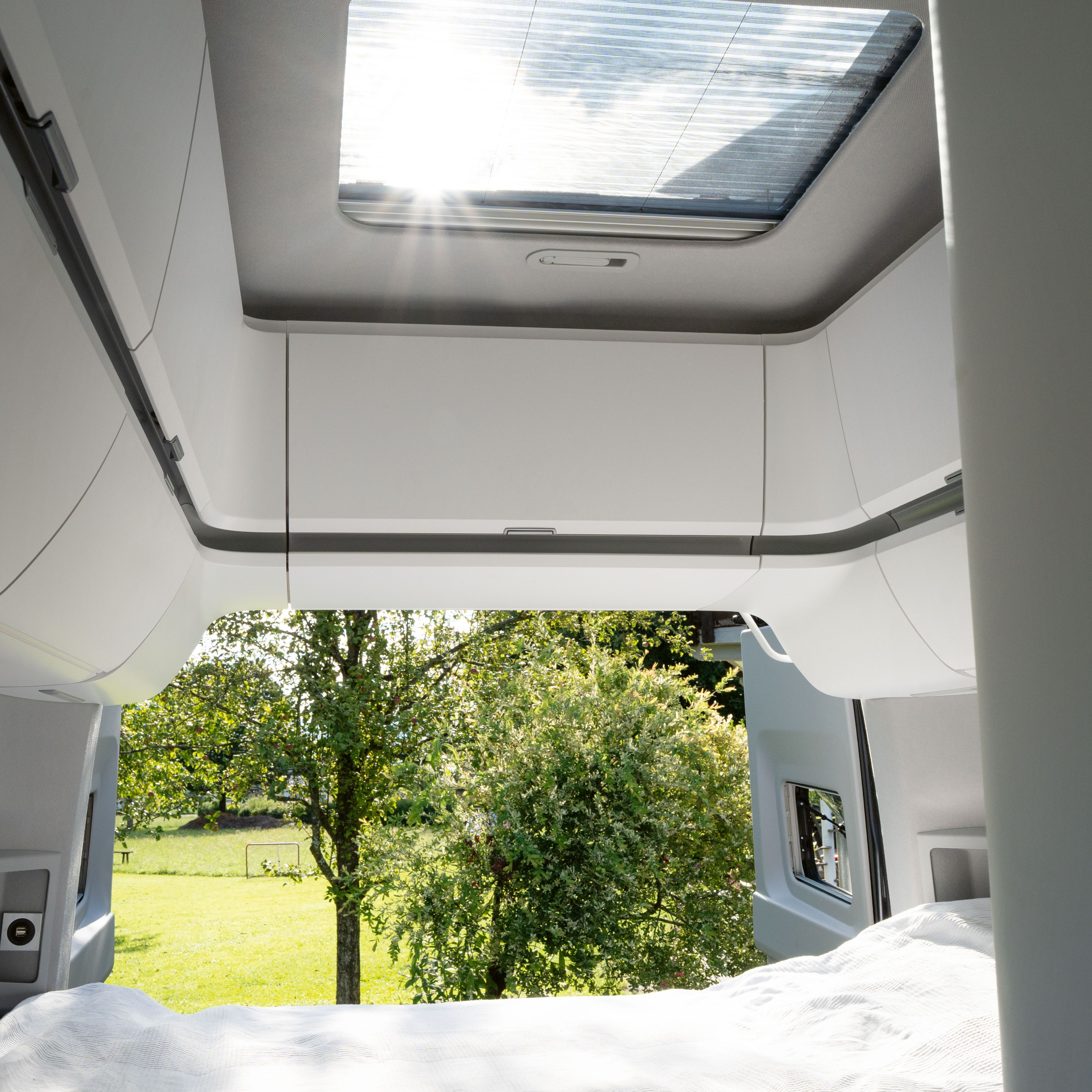Blick aus einem Wohnmobil mit einem Bett im Vordergrund und grünem Rasen und Bäumen draußen. Das Dachfenster des Fahrzeugs lässt helles Sonnenlicht herein. Links und rechts sind weiße Schränke zu erkennen.