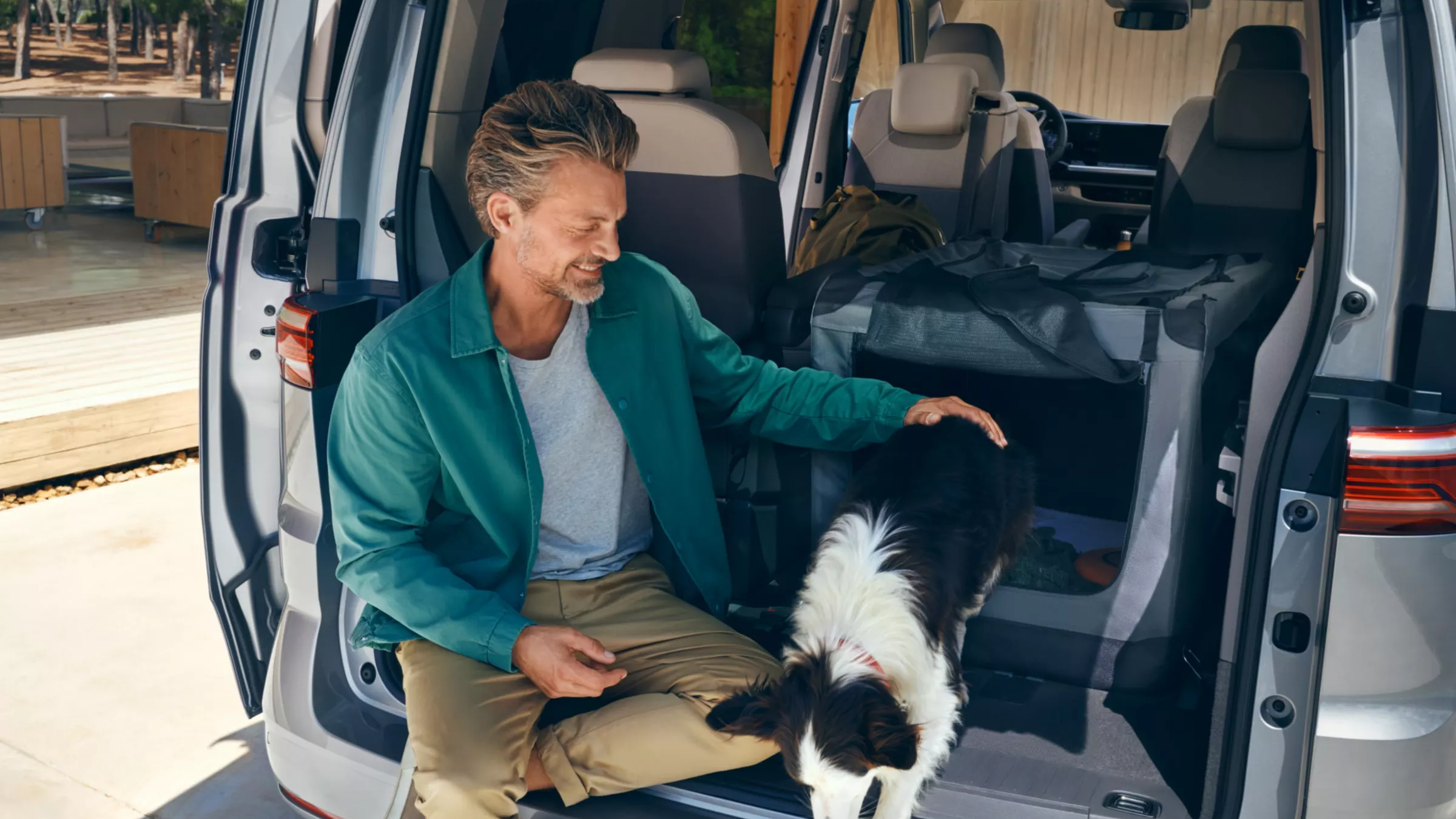 Ein Mann sitzt  im Heckbereich des neuen VW Multivans, sein Hund springt aus dem Heckbereich nach draußen