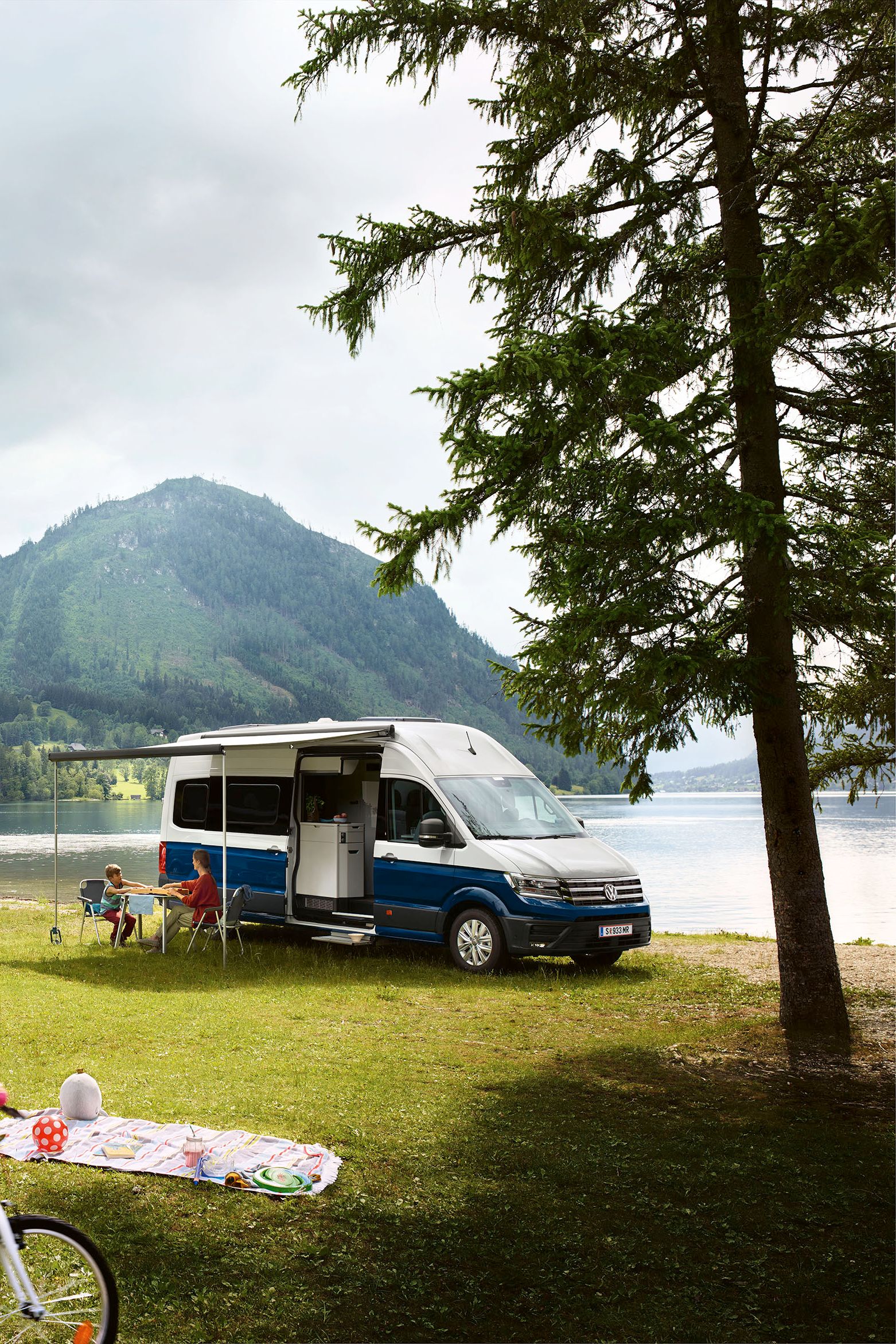 Grand California 600 е спрял край пътя, на поляна. На заден план се виждат планини и Grand California 680. 