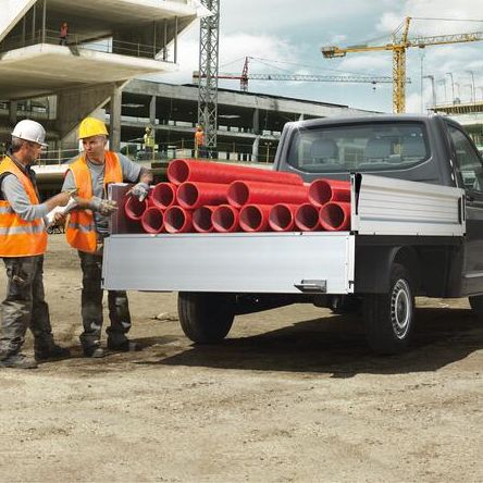 Строителна площадка с Volkswagen пикап, натоварен с червени PVC тръби, и работници с оранжеви жилетки и каски.